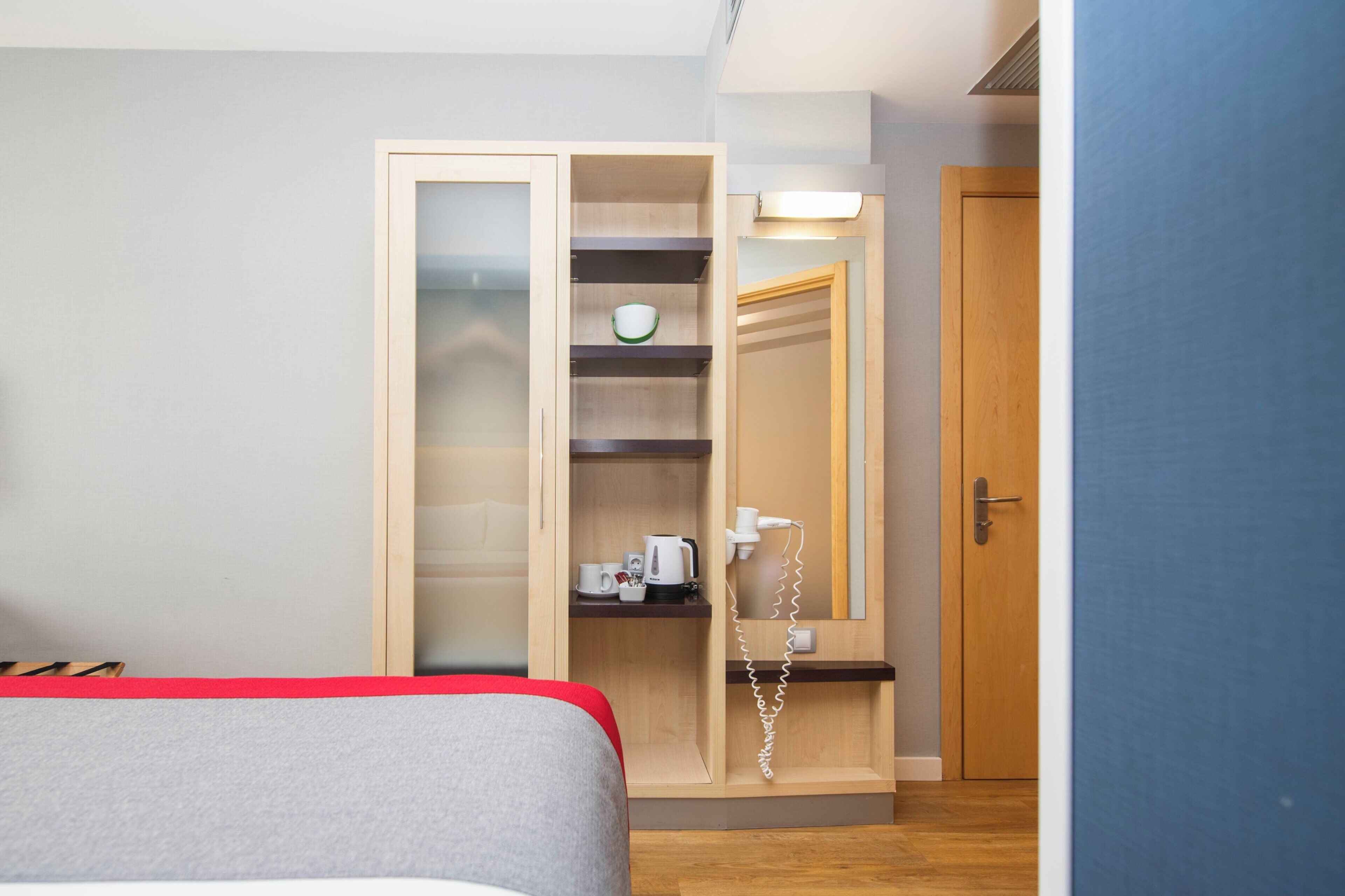 Una habitación de hotel con cama, armario con espejo, repisa con tetera y tazas, y teléfono.