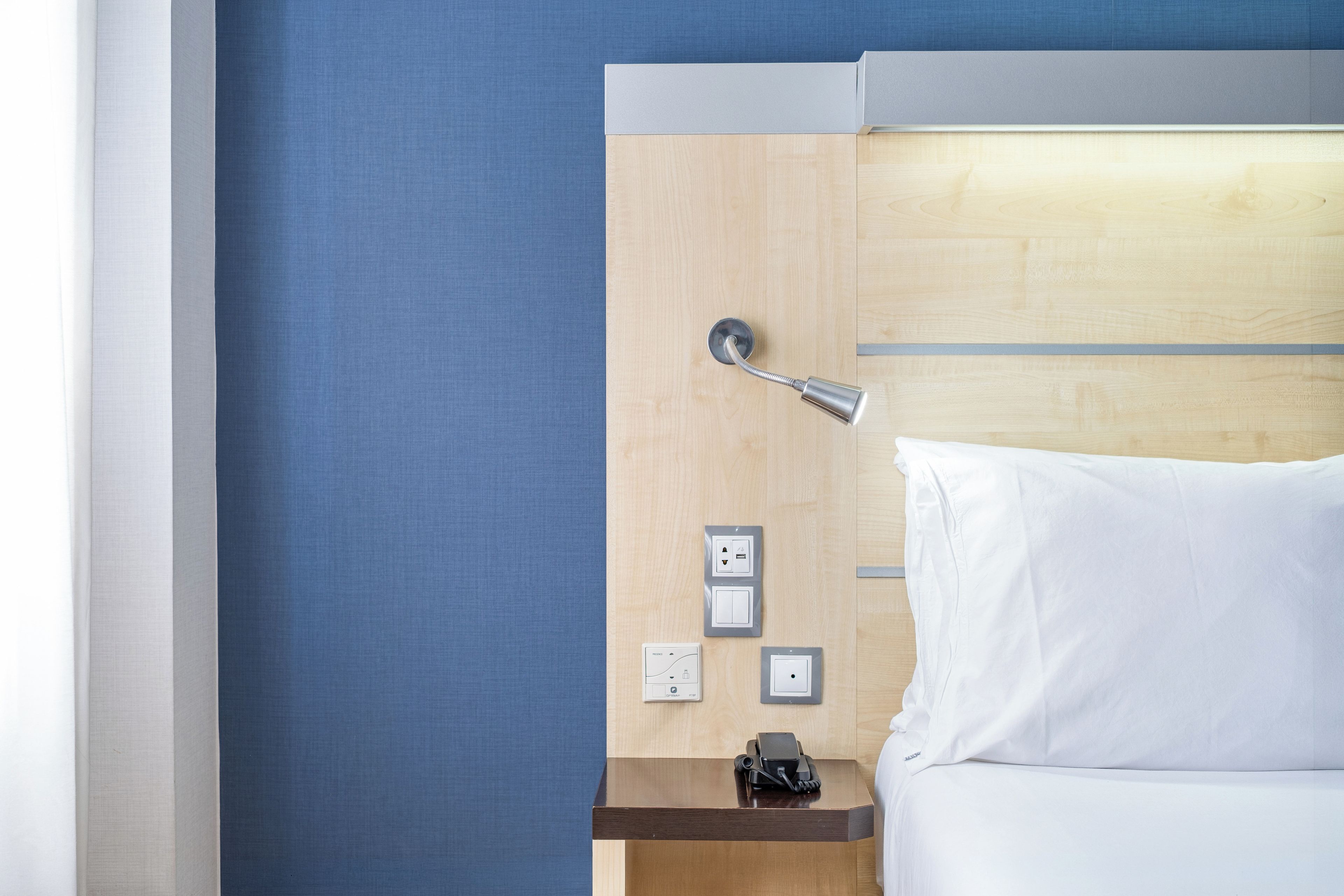 A hotel room with a wooden headboard, a bedside table with a lamp, a telephone, and a power outlet.