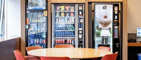A dining area with a wooden table and red chairs, a vending machine with snacks and drinks, and a view of a mountain through a glass door.