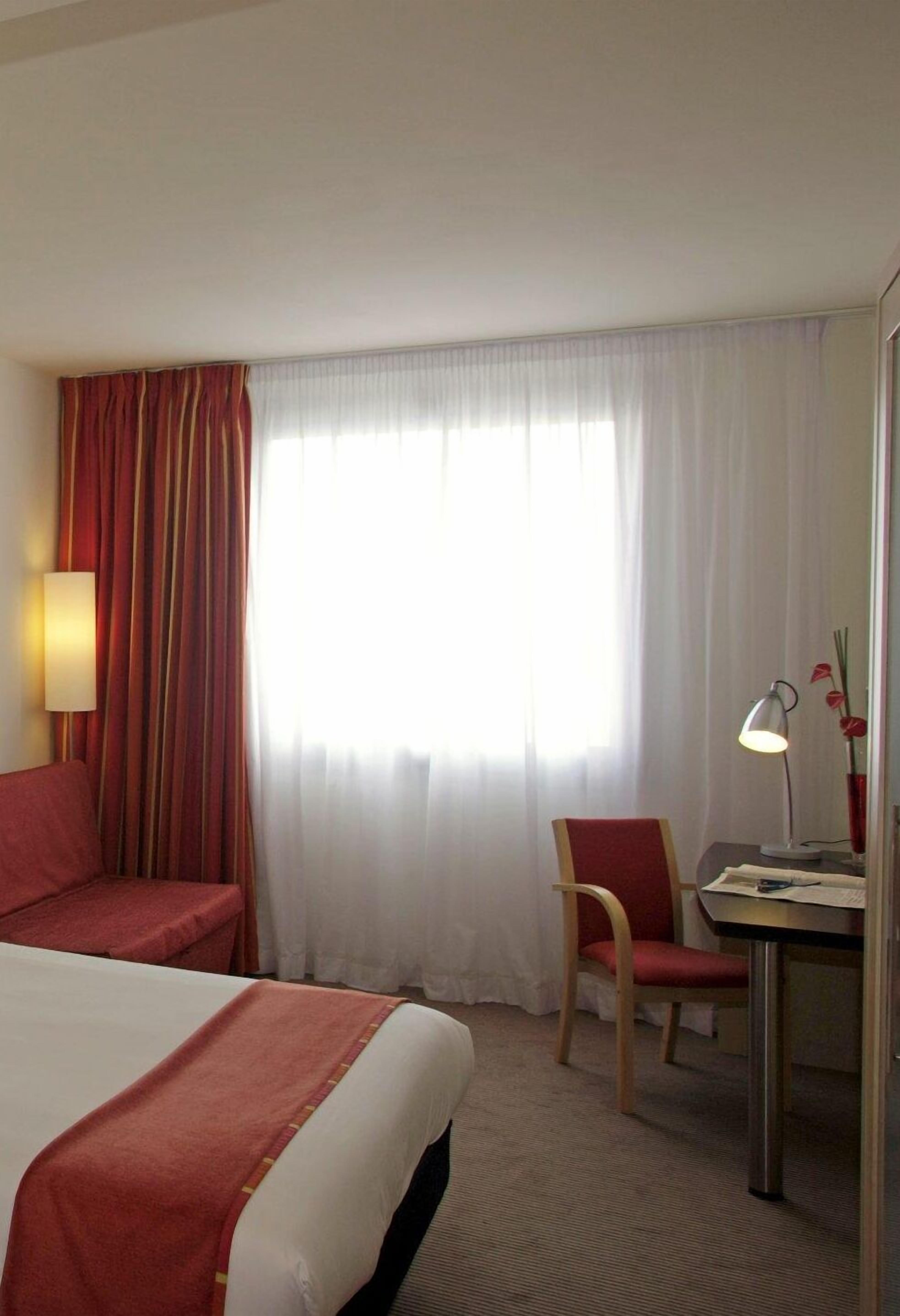 Una habitación de hotel con una cama, un escritorio con lámpara, una silla y un ventanal con cortinas.