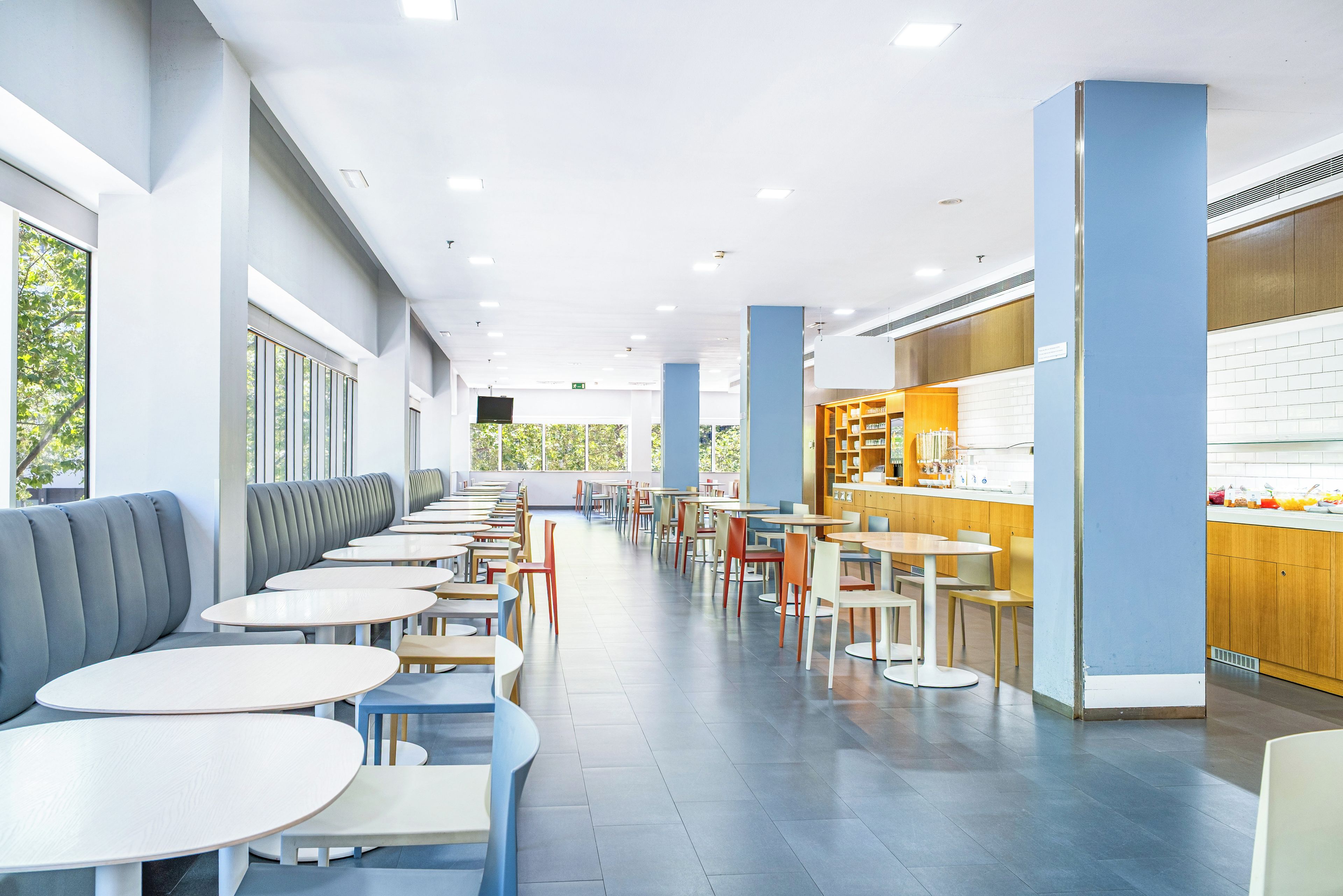 Eine moderne Cafeteria mit langen Tischen und Bänken, einem Küchenbereich und großen Fenstern.