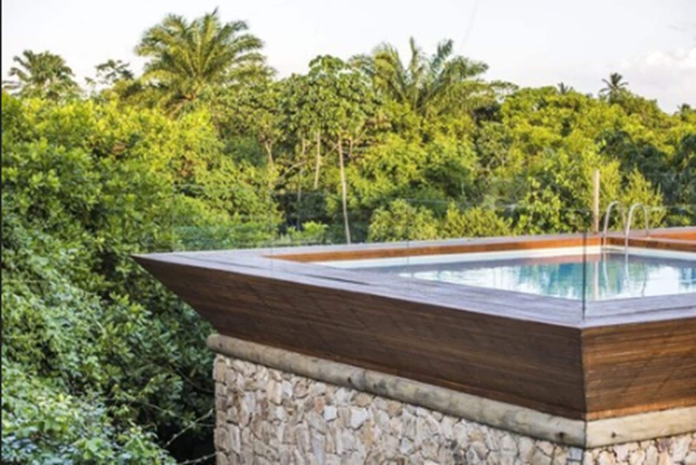 Une piscine extérieure moderne, dotée d’une terrasse en bois et de murs en pierre, entourée d’une végétation luxuriante et de palmiers.