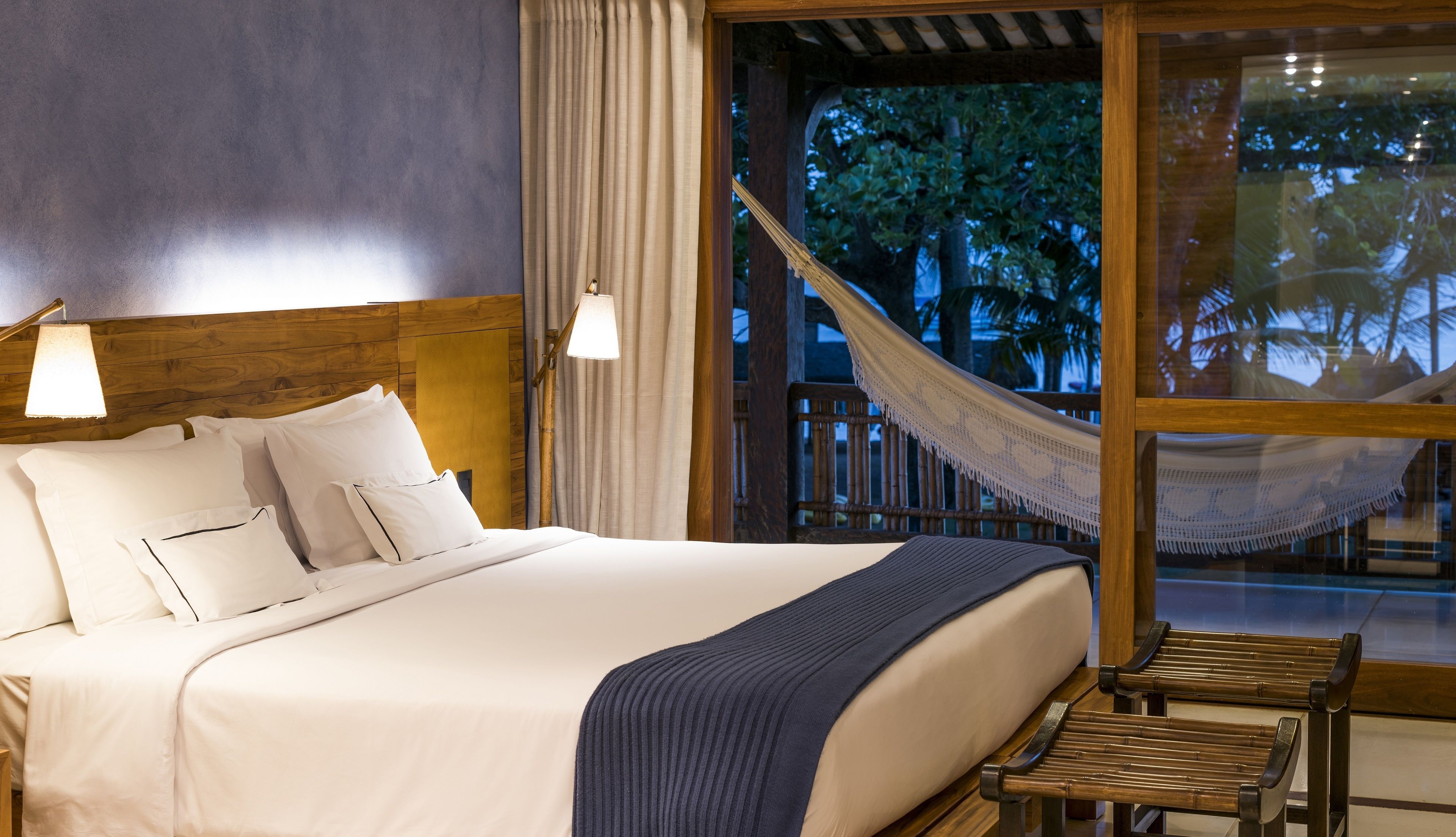 A neatly made bed with white linens and pillows, a wooden headboard, a hammock, and a view of trees through a glass door.