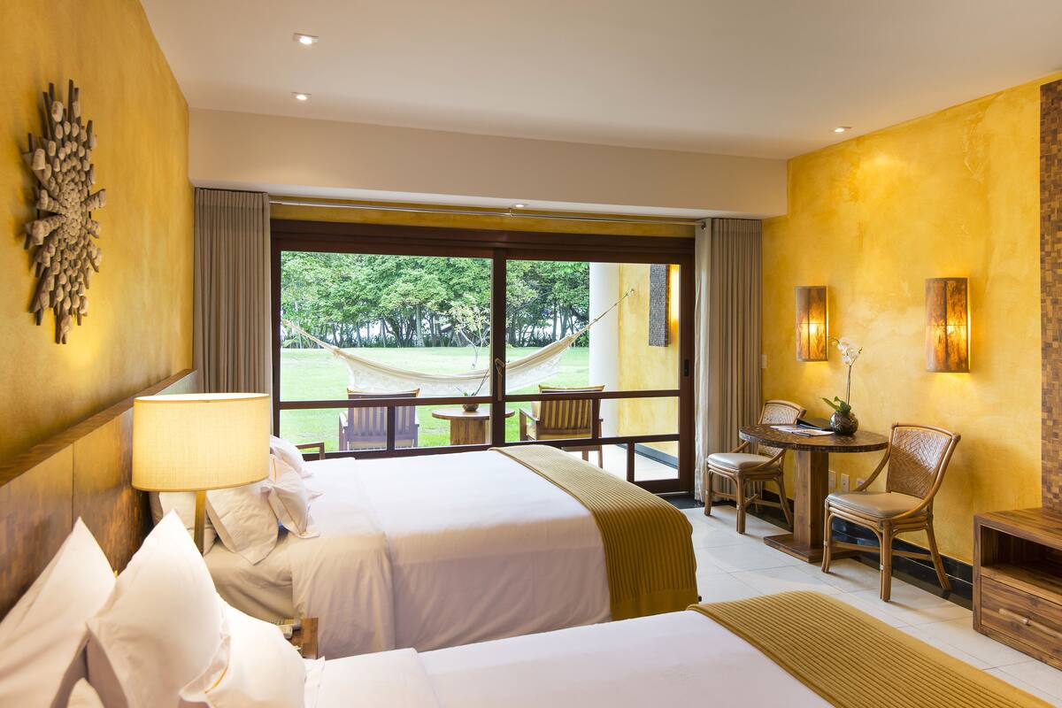A hotel room with two beds, a dining area with a table and chairs, a hammock outside, and a view of greenery.