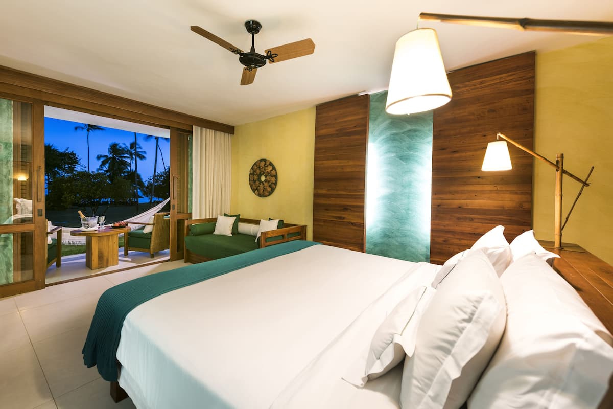 A hotel room with a large bed, a ceiling fan, a view of palm trees, and a hammock outside.