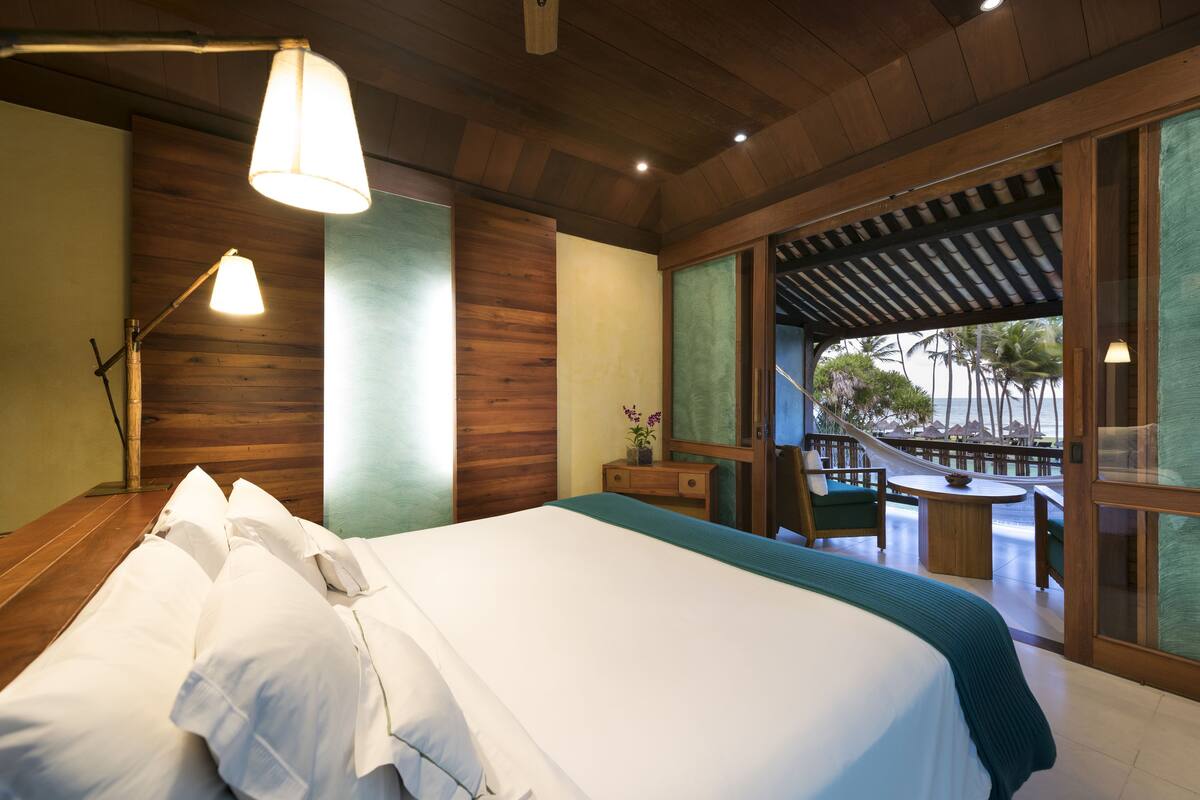 A bedroom with a large bed, a nightstand, a desk, and a view of the beach through a large window.