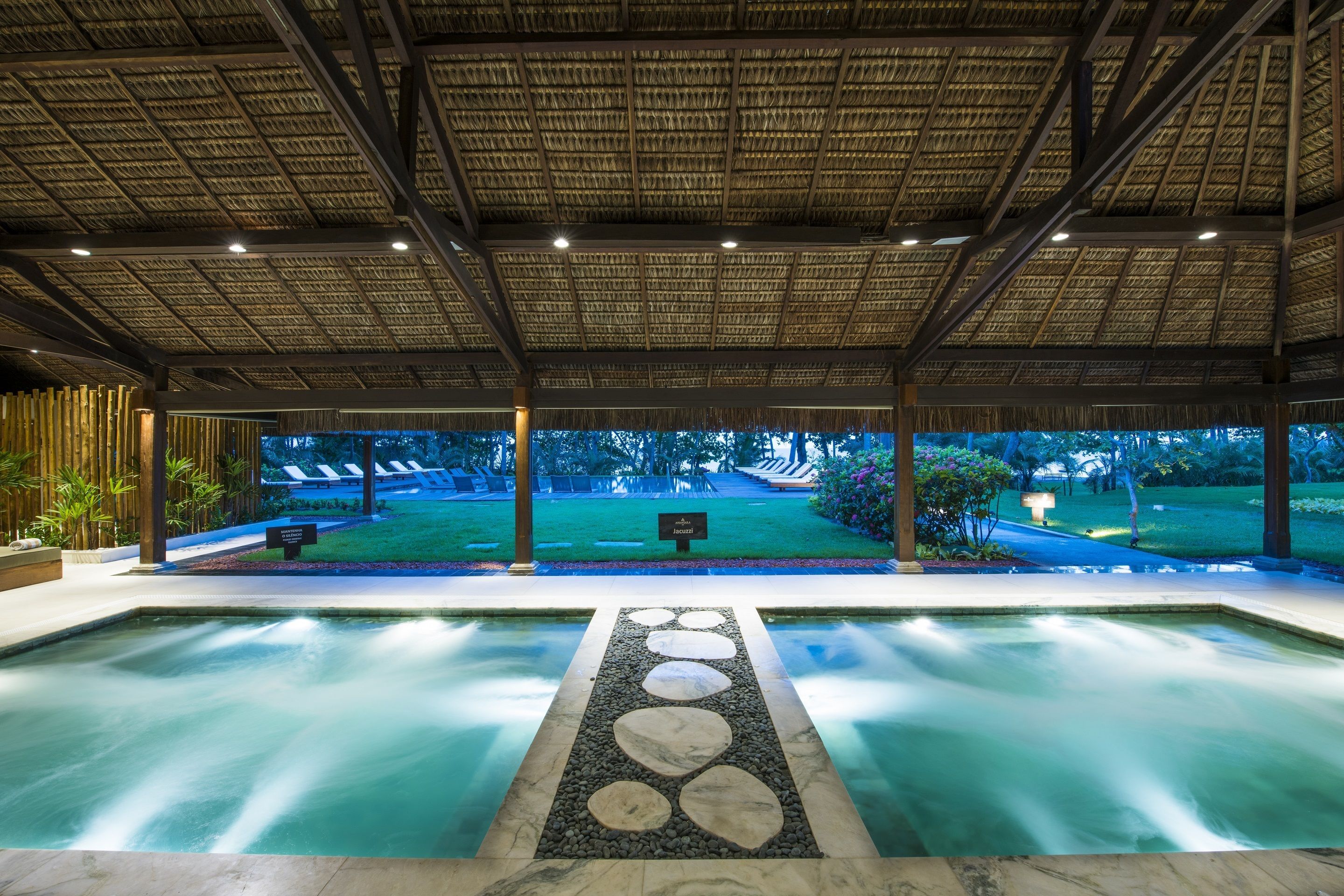 A pool area with a thatched roof, a stone pathway leading to the pool, and a view of a lawn and trees in the background.