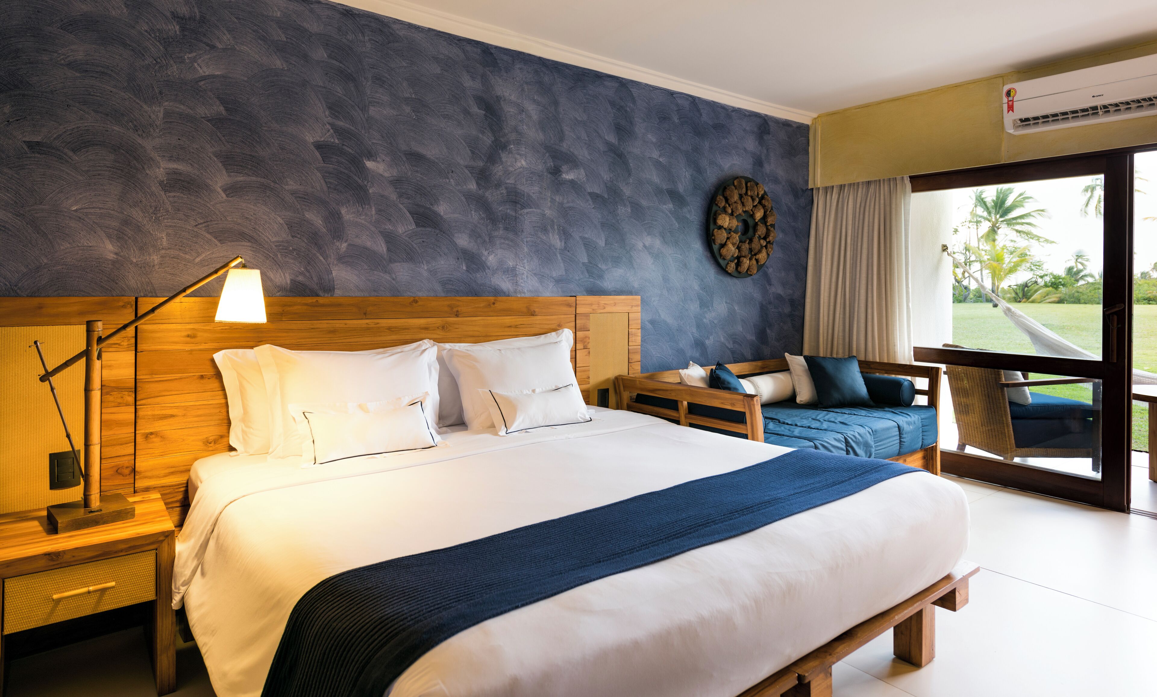 Ein Schlafzimmer mit einem großen Bett, Nachttischen, einem kleinen Tisch daneben, einem hölzernen Kopfteil, einer Wanduhr und Blick ins Freie.