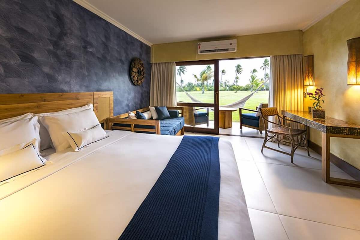 A hotel room with a large bed, a wooden headboard, a small sofa, a desk, and a view of a grassy area with palm trees.