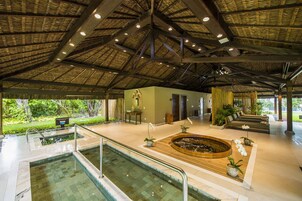 Amplia zona de piscina interior con techo de paja, jacuzzi y área de descanso con vistas al exterior.