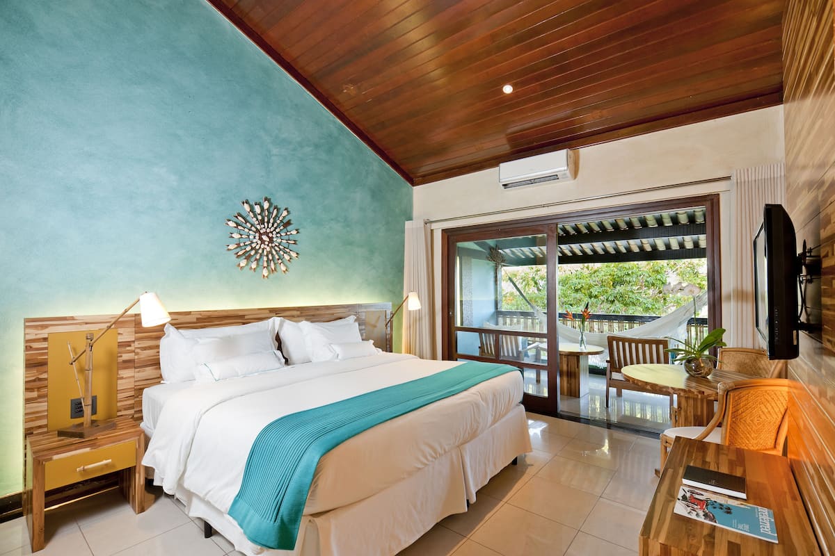 A hotel room with a large bed, wooden headboard, a nightstand with a lamp, a TV, and a balcony with a table and chairs.