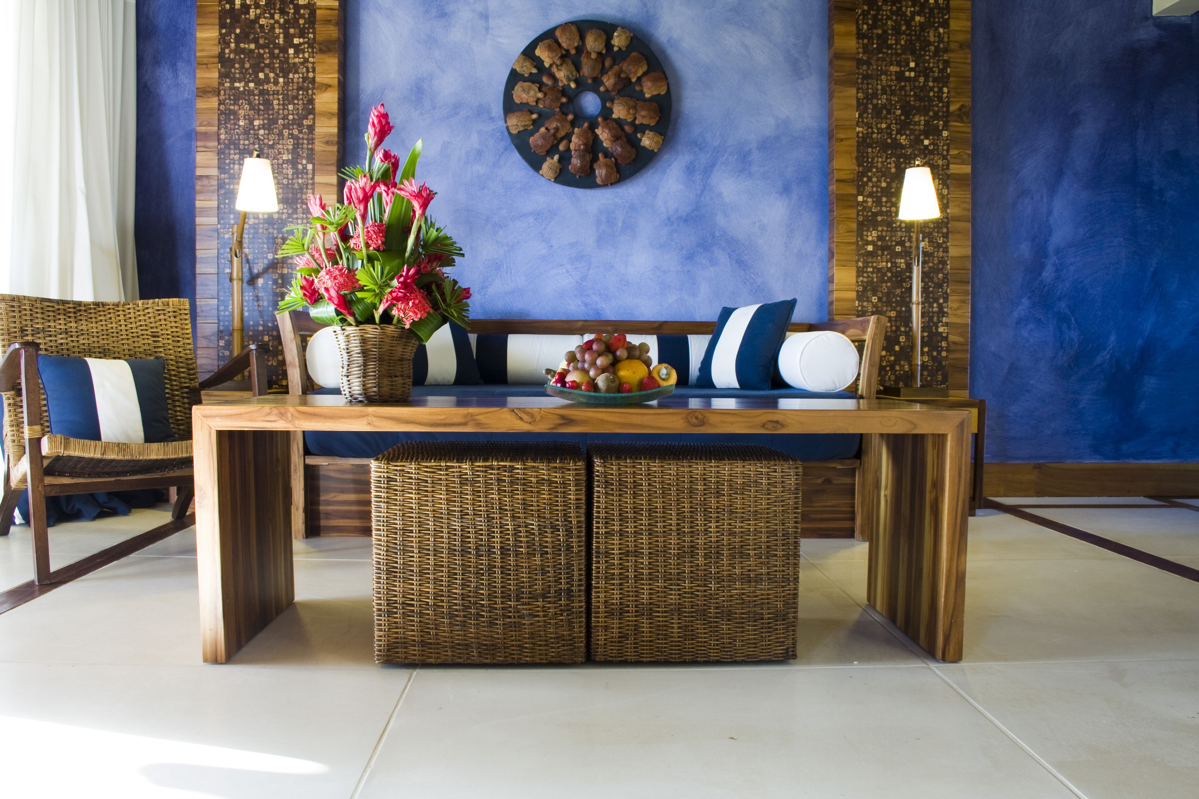 Une pièce comprenant une table à manger en bois, des chaises en osier, un canapé avec des coussins bleus et blancs, une assiette décorative accrochée au mur et deux lampadaires.