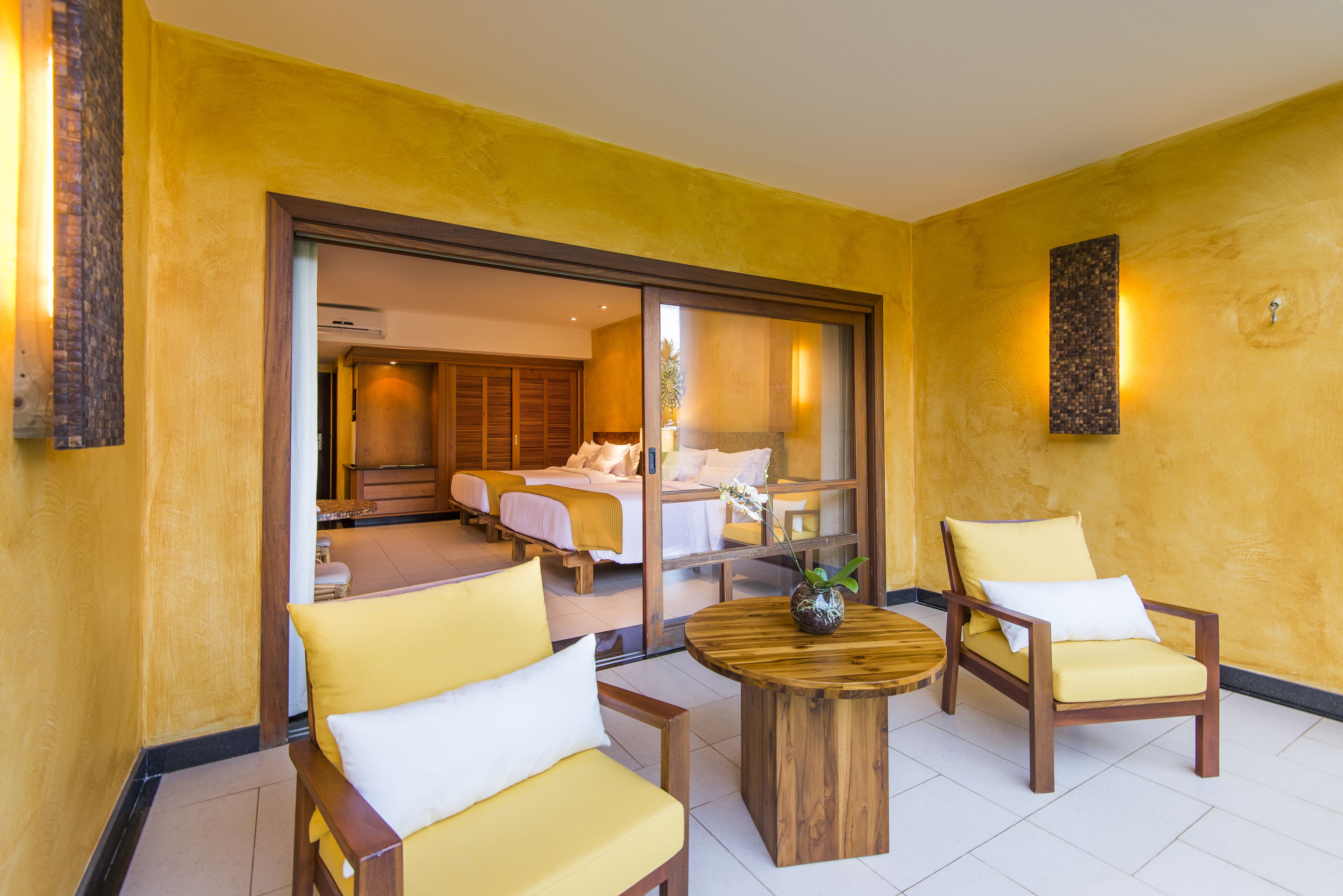 A hotel room with a yellow wall, wooden furniture, and a large sliding door leading to a bedroom with two beds.