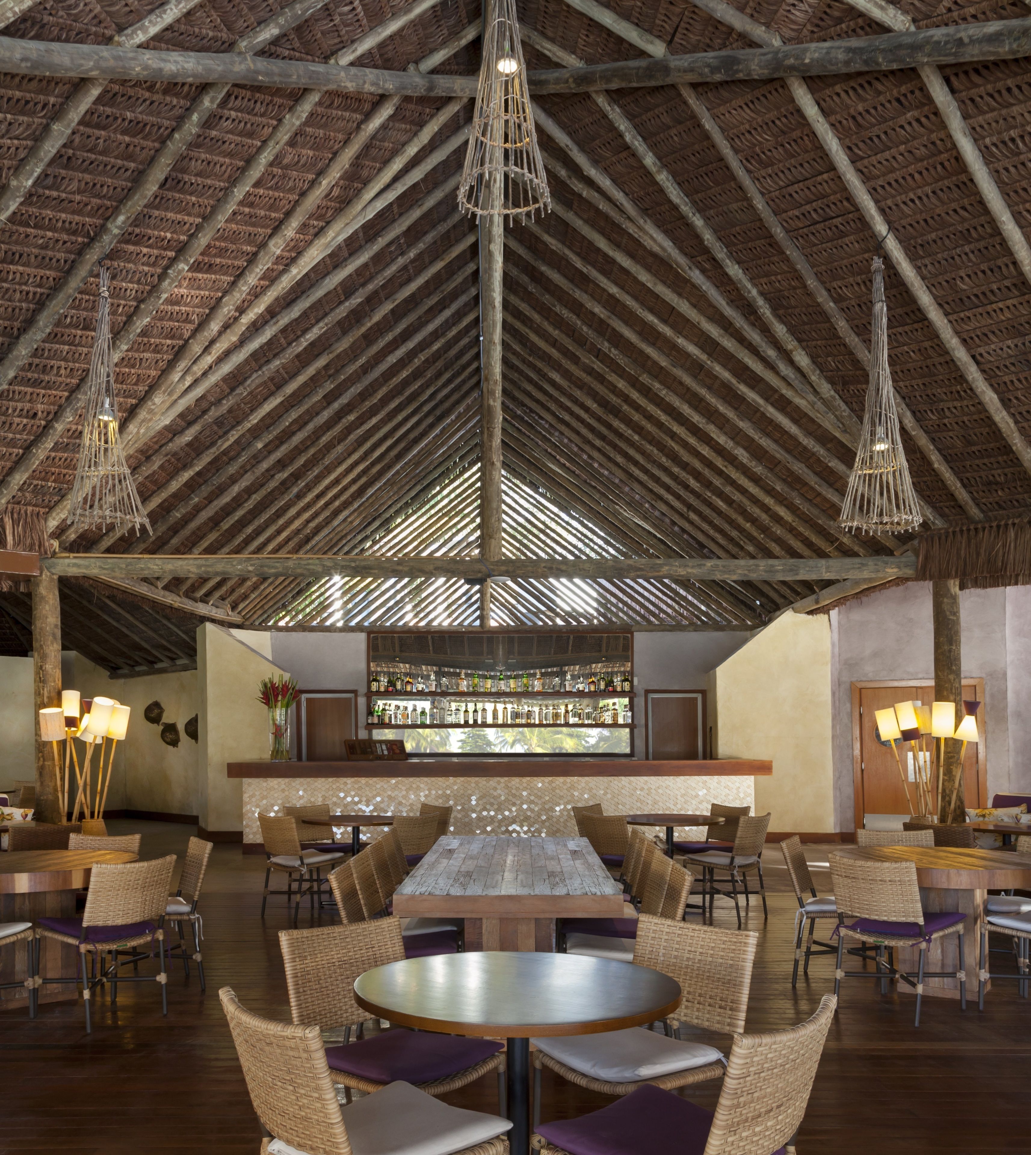A spacious indoor area with high ceilings and exposed wooden beams, featuring a bar area with a variety of bottles and a seating area with wicker furniture.