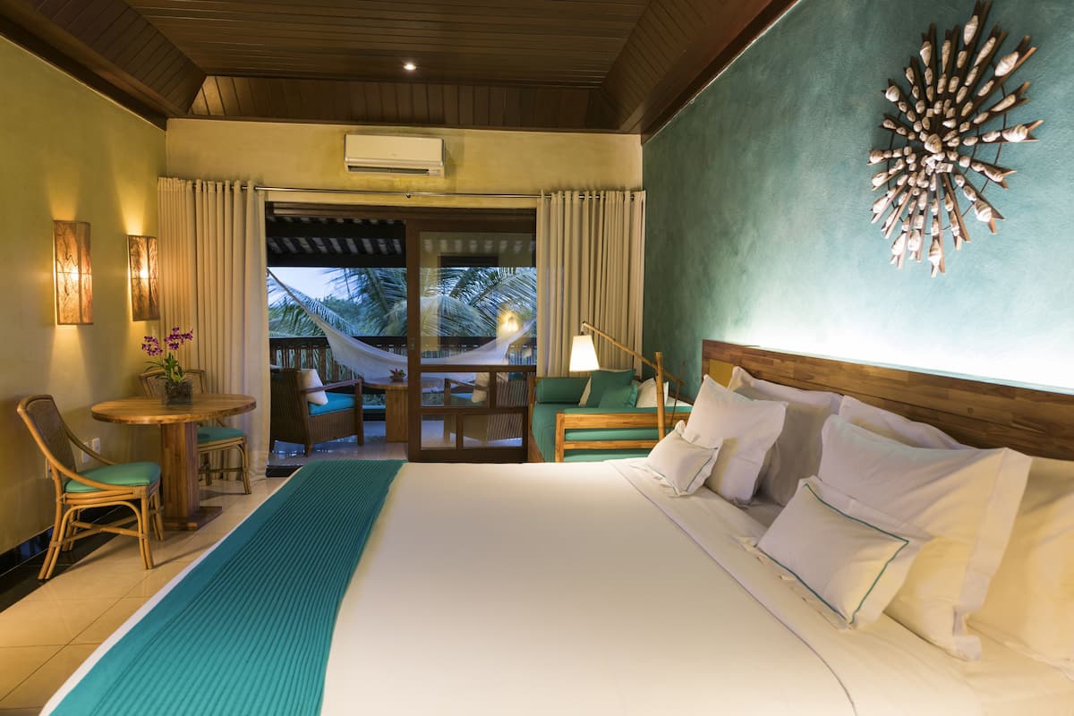 A hotel room with a large bed, a small table with chairs, a hammock, and a view of palm trees.