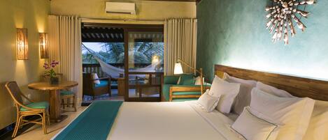 A hotel room with a large bed, a small table with chairs, a hammock, and a view of palm trees.