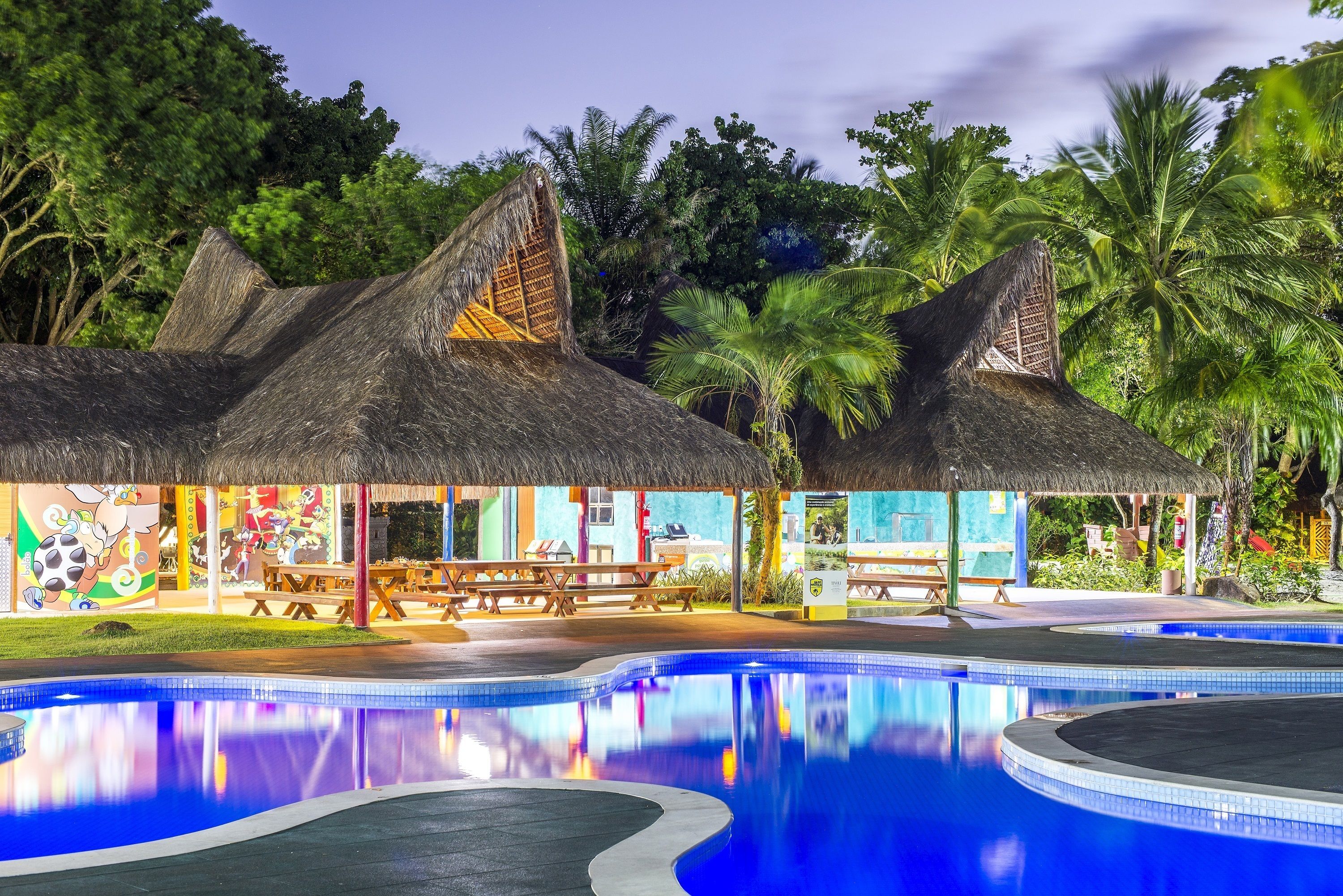 Un área de piscina con una estructura de techo de paja, zona de comedor y exuberante jardín tropical.