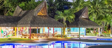 Un área de piscina con una estructura de techo de paja, zona de comedor y exuberante jardín tropical.