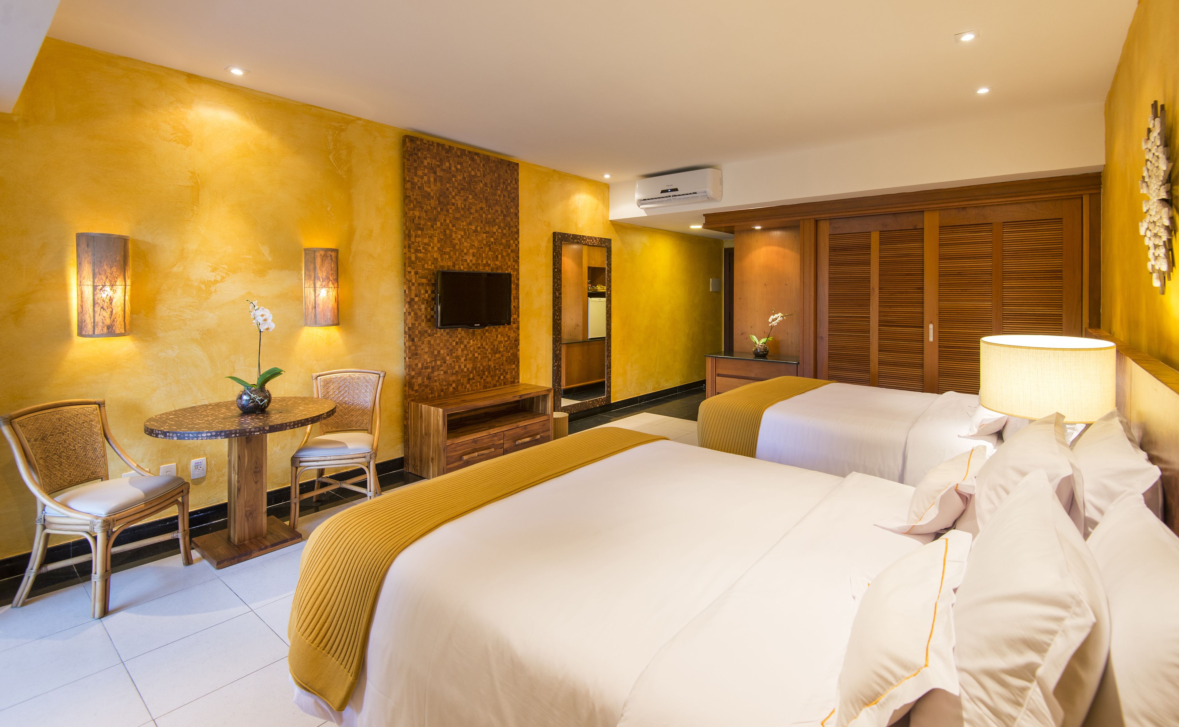 A hotel room with two beds, a seating area with a table and chairs, a TV mounted on the wall, and a wooden wardrobe.