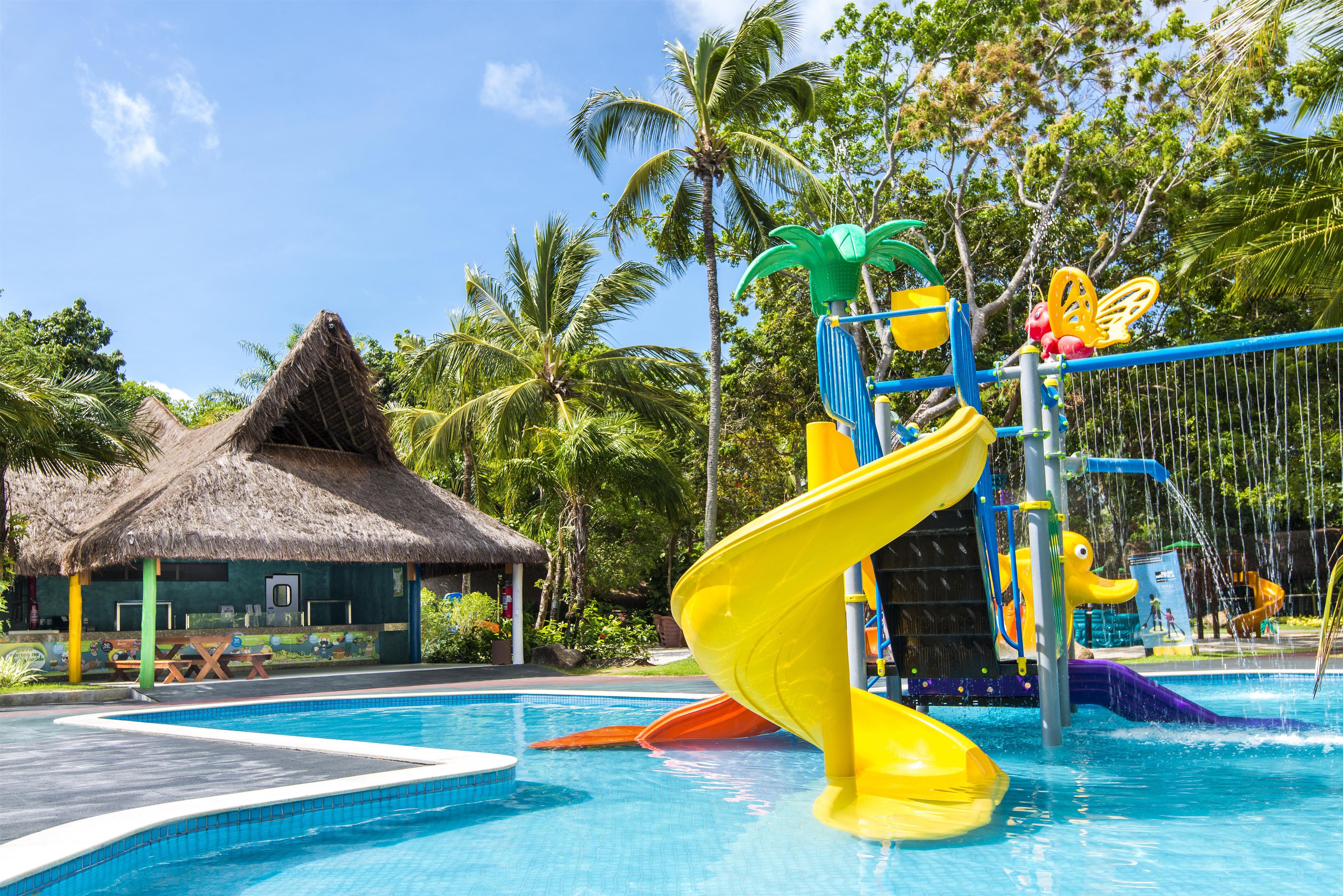 A swimming pool with a water slide, a thatched-roof hut, and palm trees.