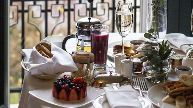 Una mesa lista para el desayuno con vista a la ciudad, que incluye un jarrón con flores, una cafetera y diferentes productos para el desayuno.