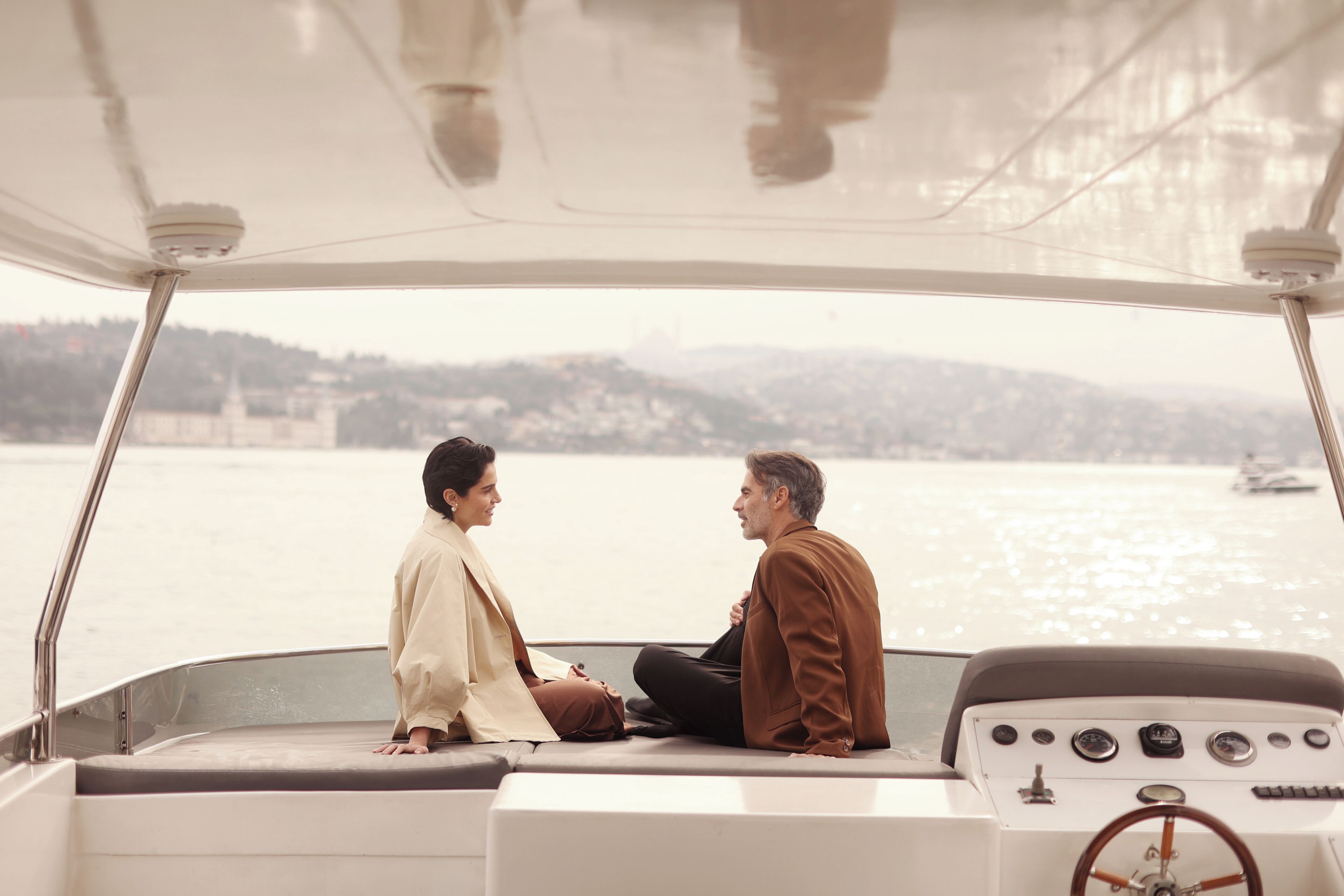L'intérieur d'un bateau avec un volant, deux personnes assises face à face, et une vue sur une étendue d'eau avec de la terre ferme au loin.
