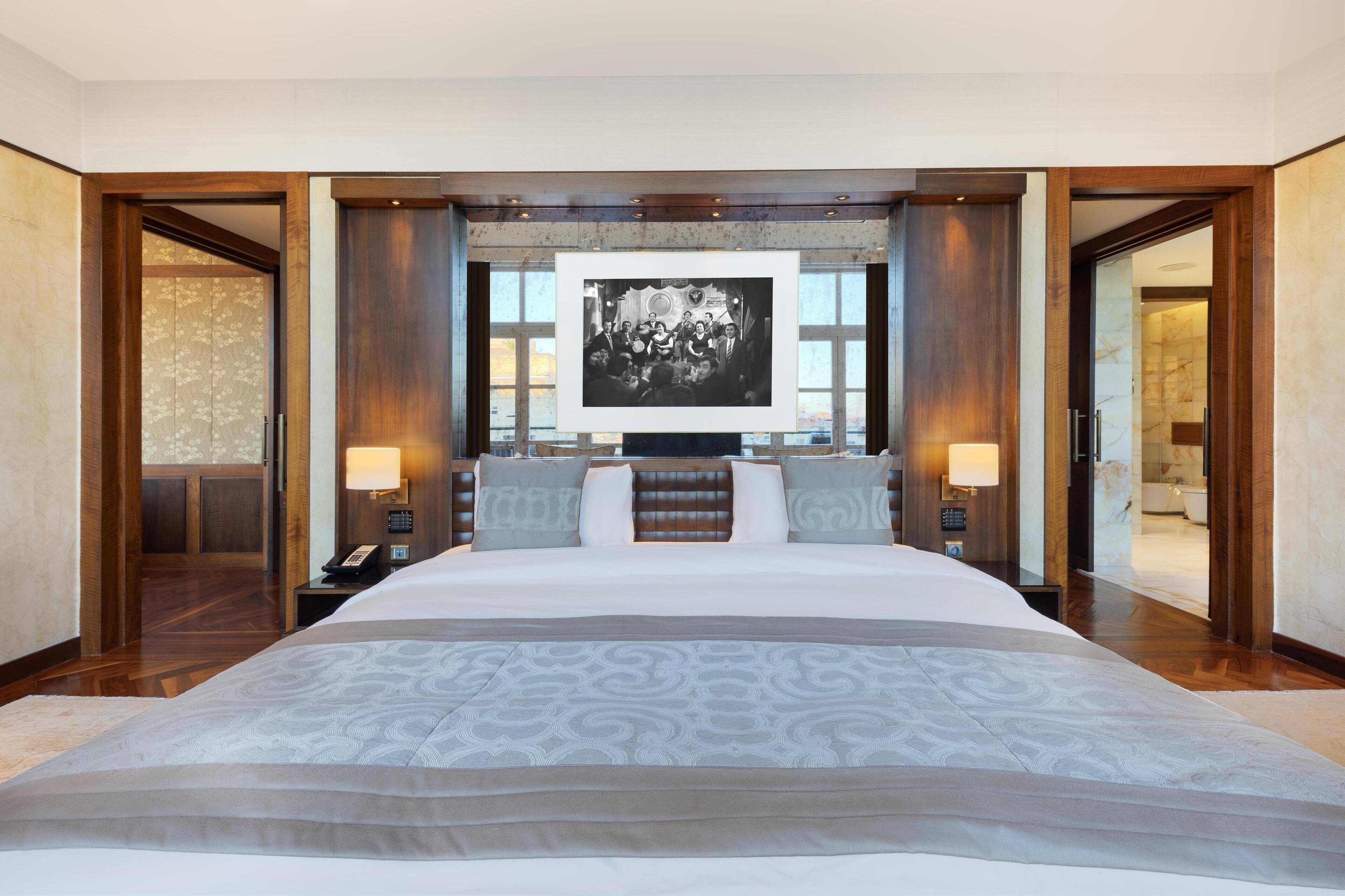 A large bed with a patterned bedspread, two bedside tables with lamps, a large framed picture above the bed, and wooden paneling on the walls.