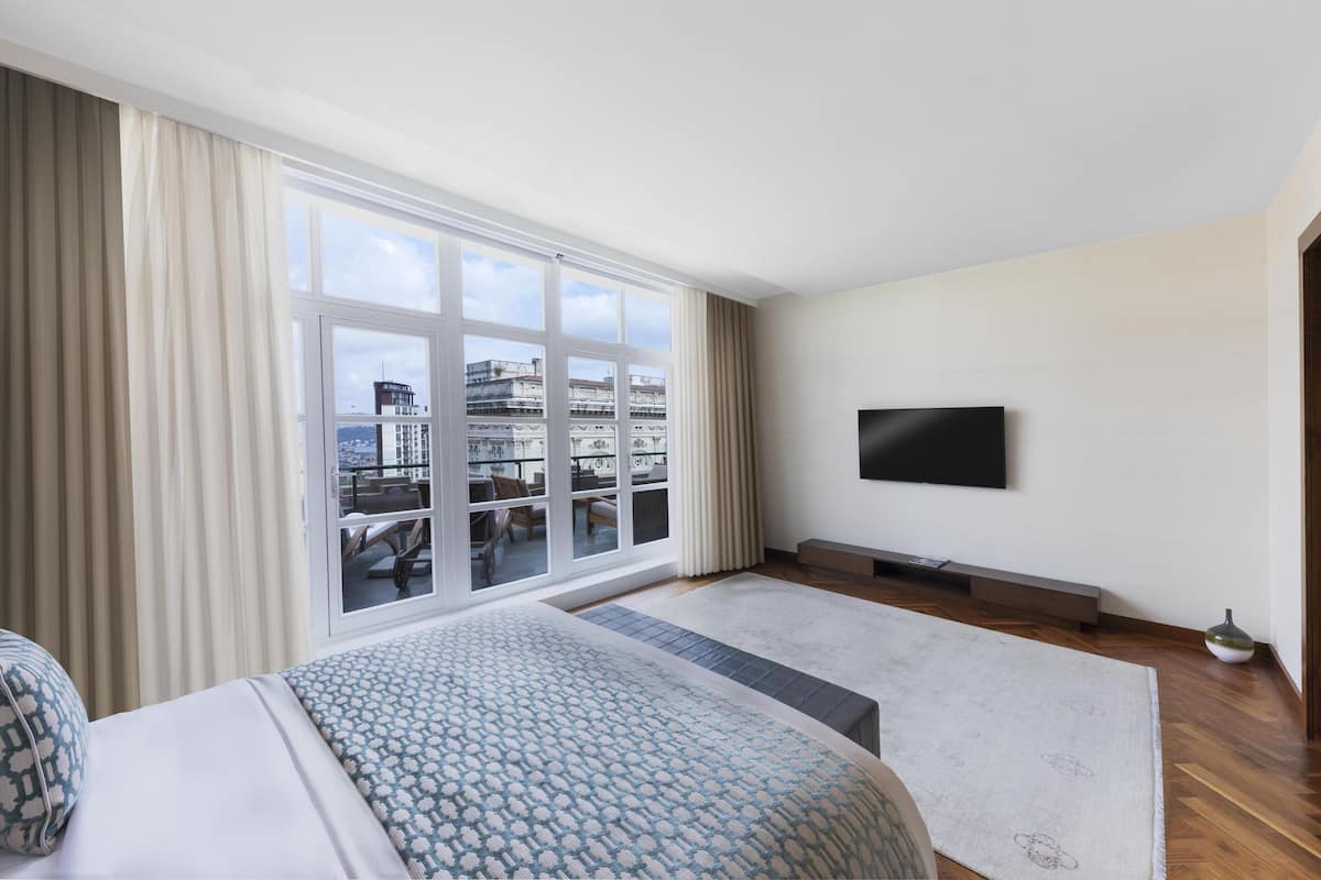 Habitación de hotel con una ventana grande, una cama, un televisor y vistas a edificios.