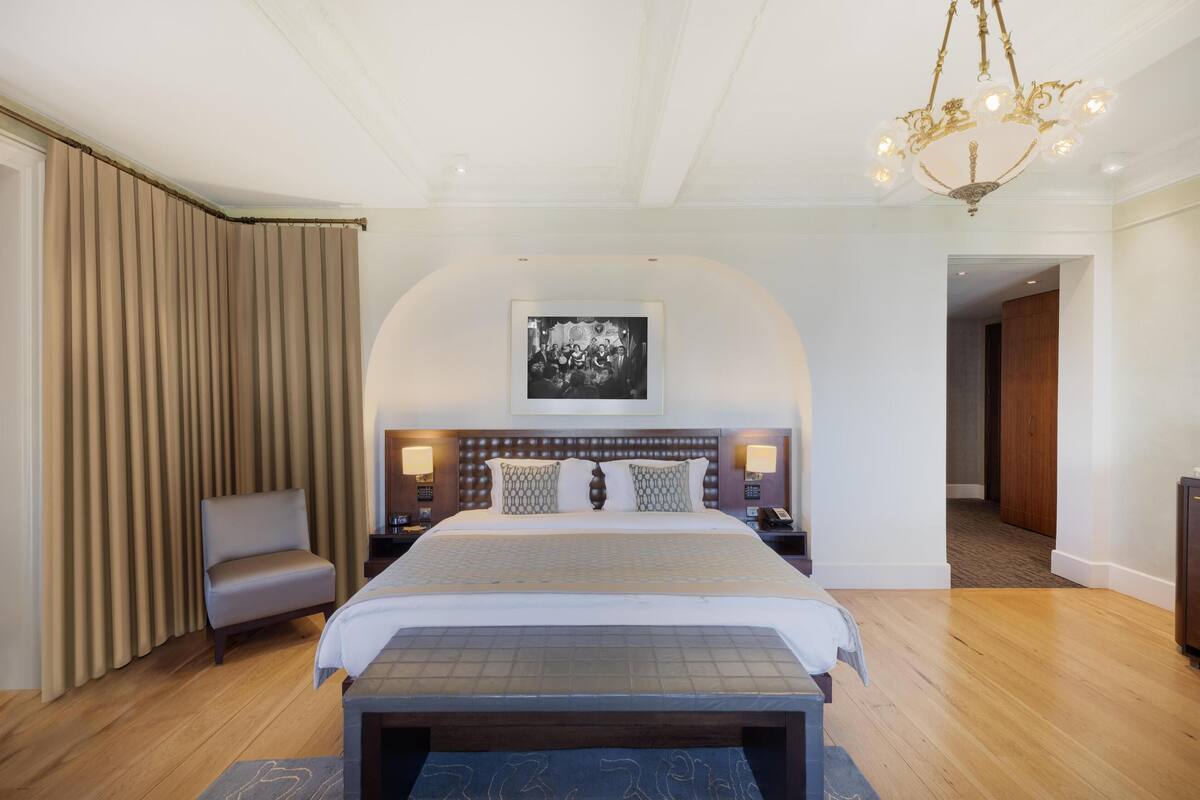 Habitación de hotel con una cama grande, un banco al pie de la cama, una silla, una mesita de noche con una lámpara y un candelabro.