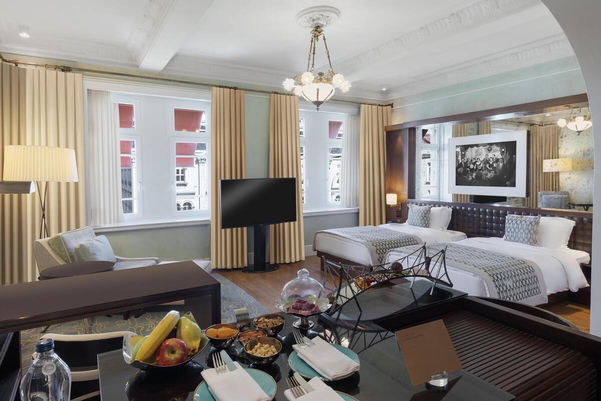 Habitación de hotel con cama, televisión, una mesa de comedor con fruta y una botella, y un candelabro.