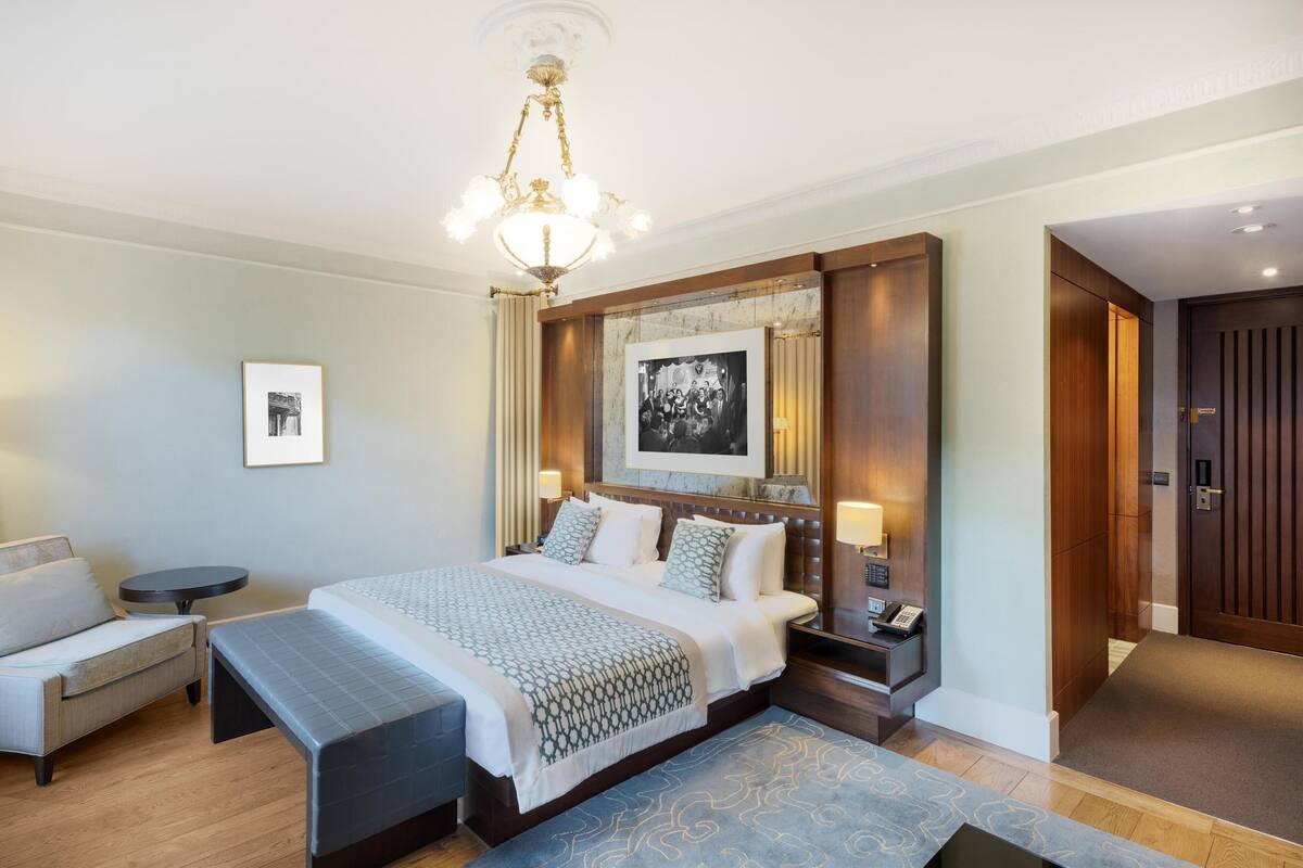 Una habitación de hotel con una cama grande, mesitas de noche, un banco, un escritorio y una araña.