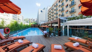 Zona de piscina en la azotea con tumbonas de madera y sombrillas naranjas, rodeada de edificios.