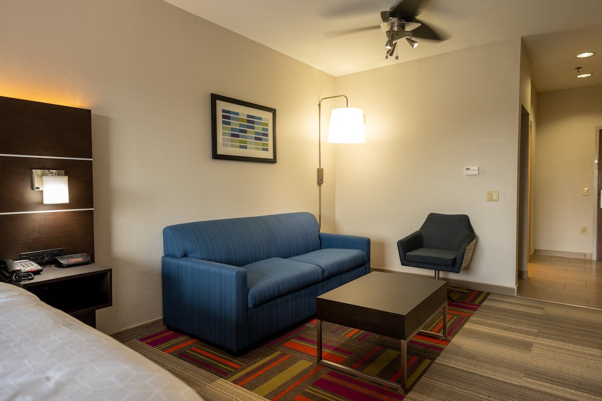 A hotel room with a blue sofa, a small armchair, a coffee table, a lamp, a framed picture on the wall, and a bed.