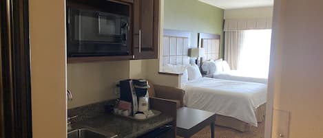 Habitación de hotel compacta con kitchenette, que incluye microondas, cafetera y pileta. La habitación tiene una cama con ropa blanca y una ventana con cortinas.