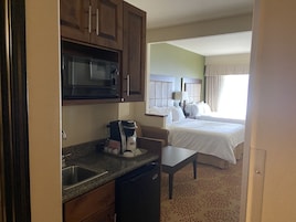 Quarto de hotel compacto com cozinha americana, incluindo micro-ondas, máquina de café e pia. O quarto possui cama com roupa de cama branca e janela com cortinas.