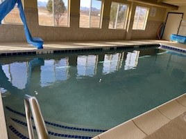 Piscina interna com água cristalina, um escorregador azul e vista para o exterior através de grandes janelas.