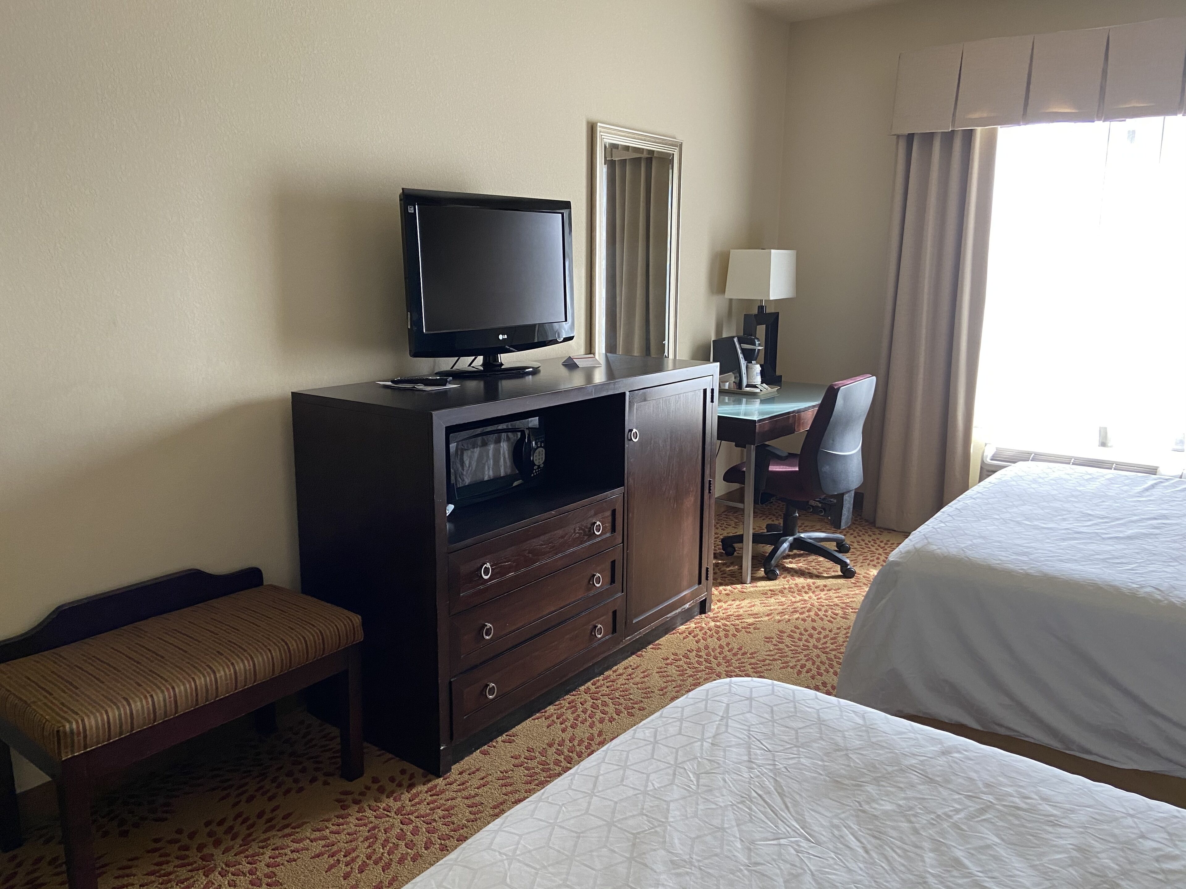 A hotel room with a bed, a desk with a chair, a television on a dresser, and a window with curtains.