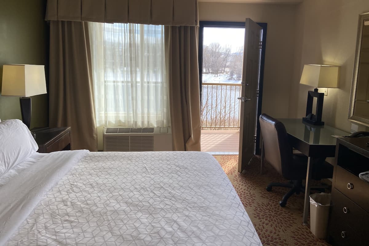 Ein Hotelzimmer mit einem großen Bett, einem Schreibtisch, einem Sessel und Blick auf eine verschneite Landschaft durch eine offene Tür.