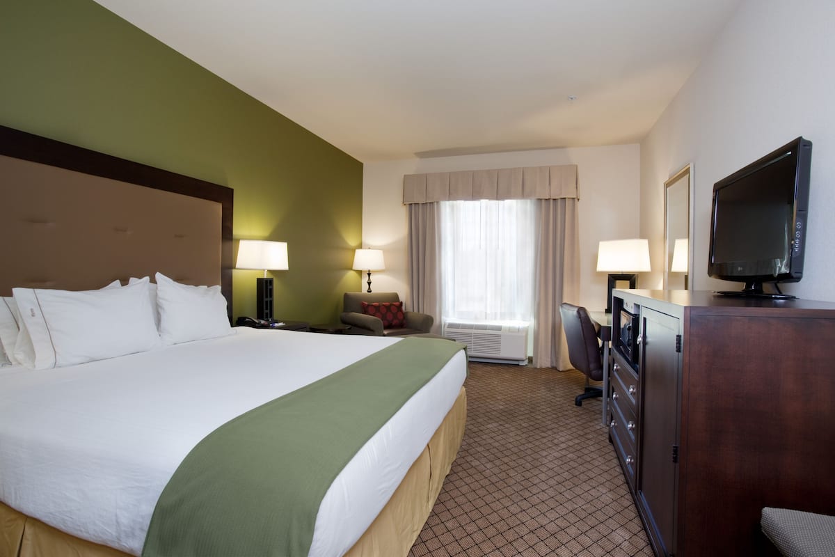 Ein Hotelzimmer mit einem großen Bett, zwei Nachttischlampen, einem Schreibtisch mit Computer, einem Fernseher und einem Fenster mit Vorhängen.