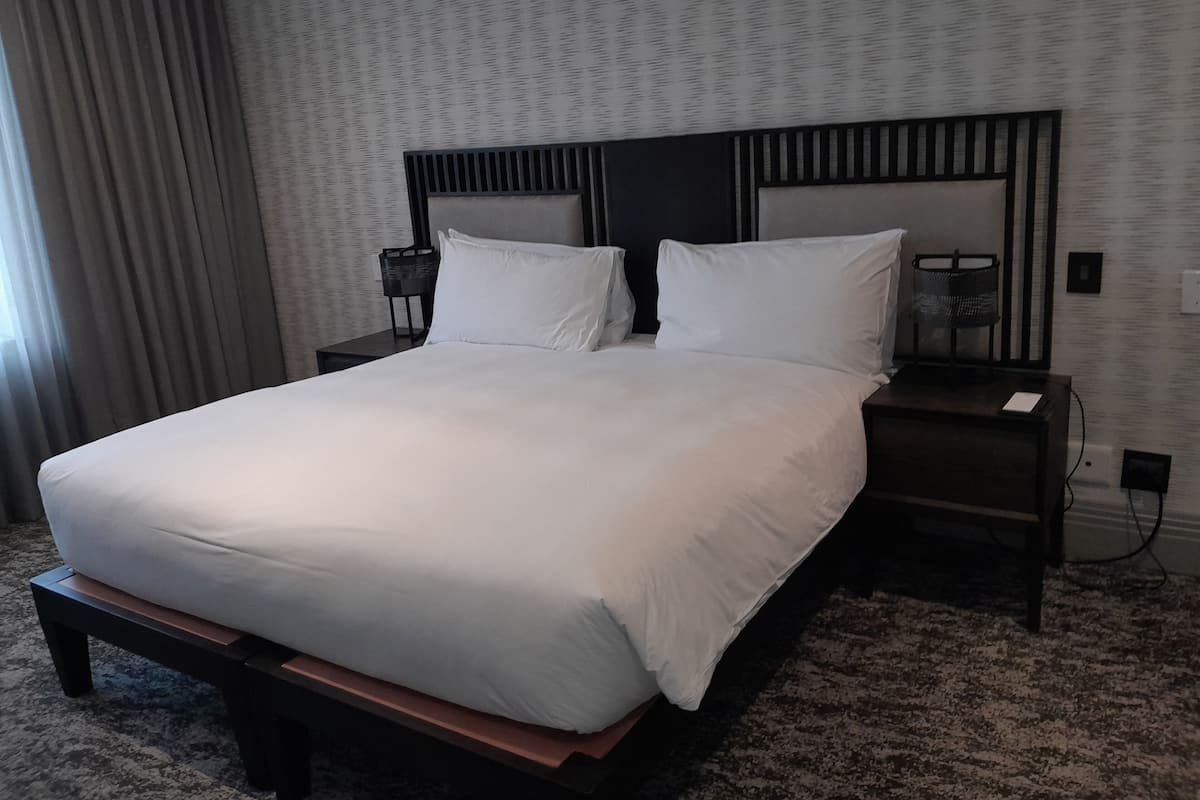 Una cama grande con ropa blanca, un cabecero oscuro y un piecero de madera. Hay dos mesitas de noche con lámparas y una pequeña mesa junto a la cama. La habitación tiene una alfombra estampada y una ventana con cortinas.