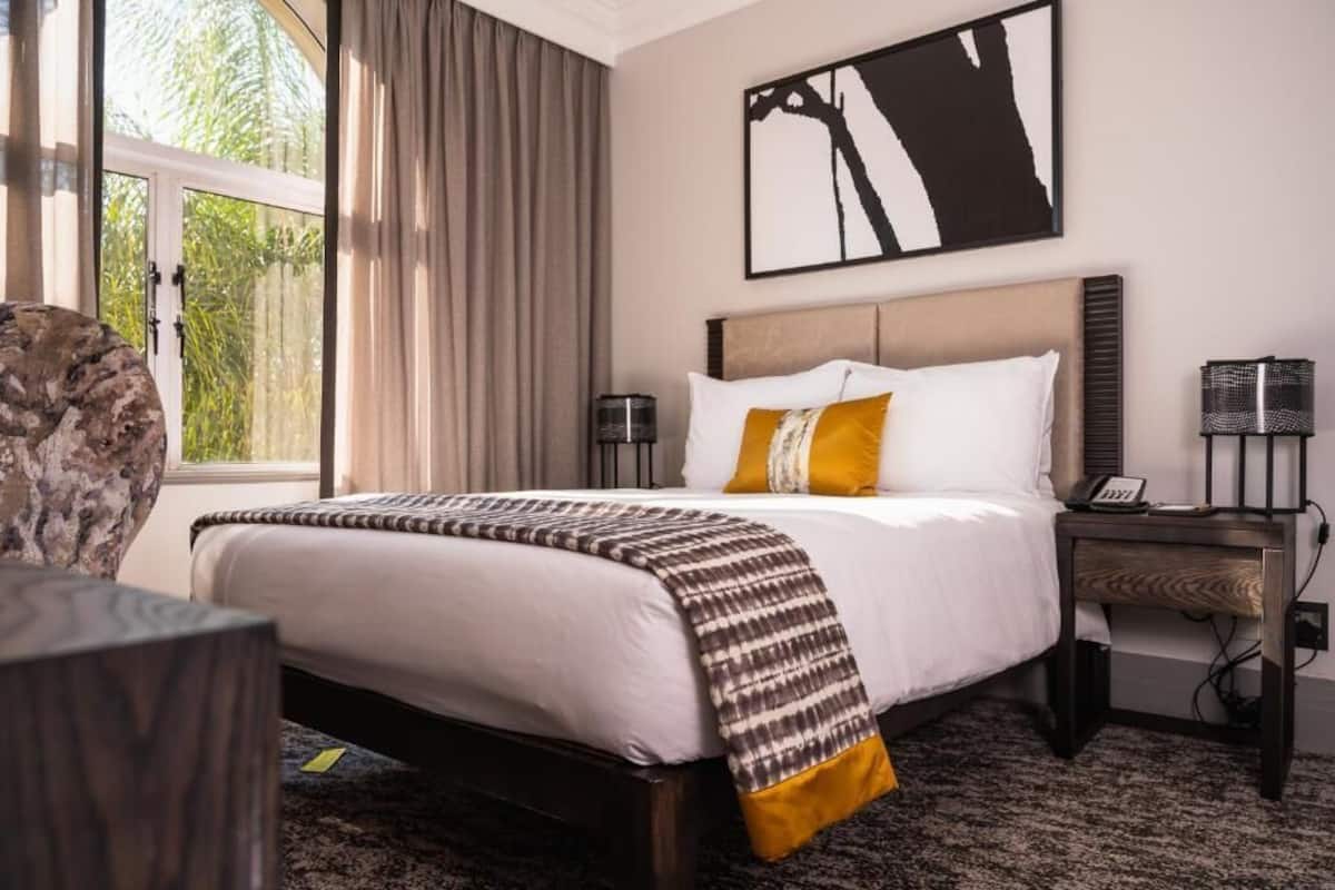 Una habitación de hotel con cama, mesita de noche, lámpara y ventana con cortinas.