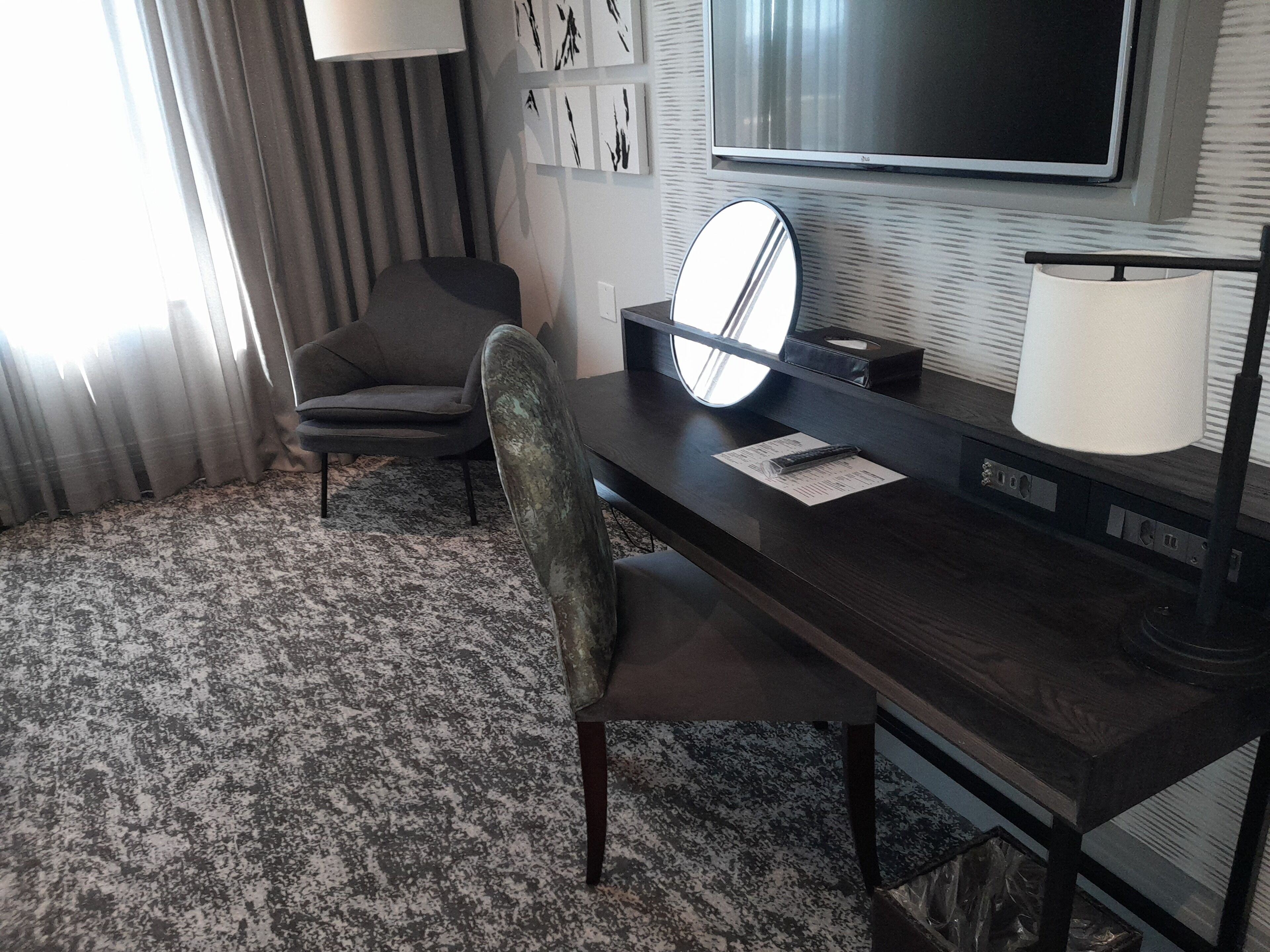 Ett hotellrum med ett mörkt skrivbord i trä, en stol, en platt-TV som är monterad på väggen, en lampa och en mönstrad matta.