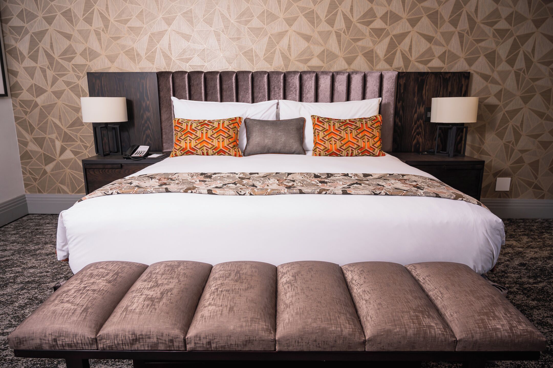 Cama bem arrumada com colcha estampada, duas luminárias de cabeceira e um banco de madeira aos pés da cama.