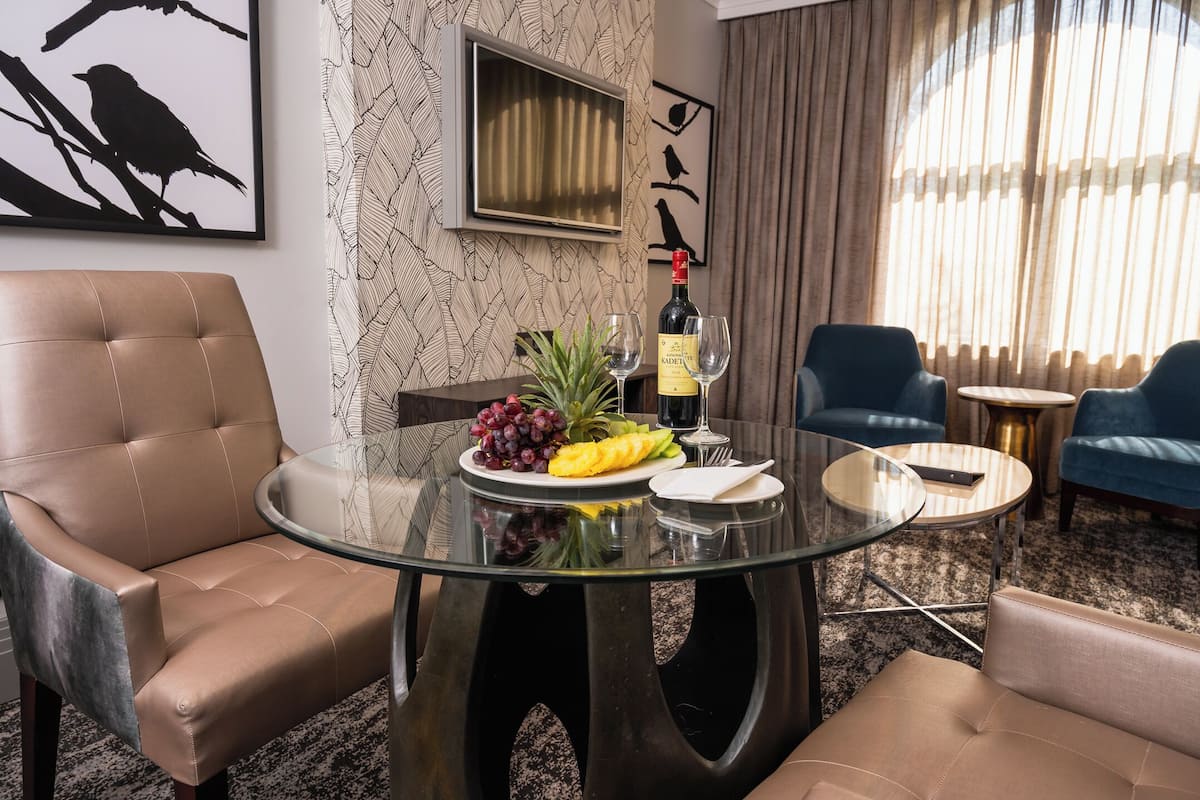 Una habitación de hotel moderna con una mesa de vidrio, dos sillones de cuero, un sofá, un televisor y un gran ventanal con cortinas.