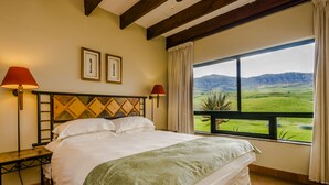 Ein Schlafzimmer mit einem großen Bett, zwei Nachttischlampen, Blick auf eine grüne Landschaft durch ein Fenster und einem Holzbalken an der Decke.