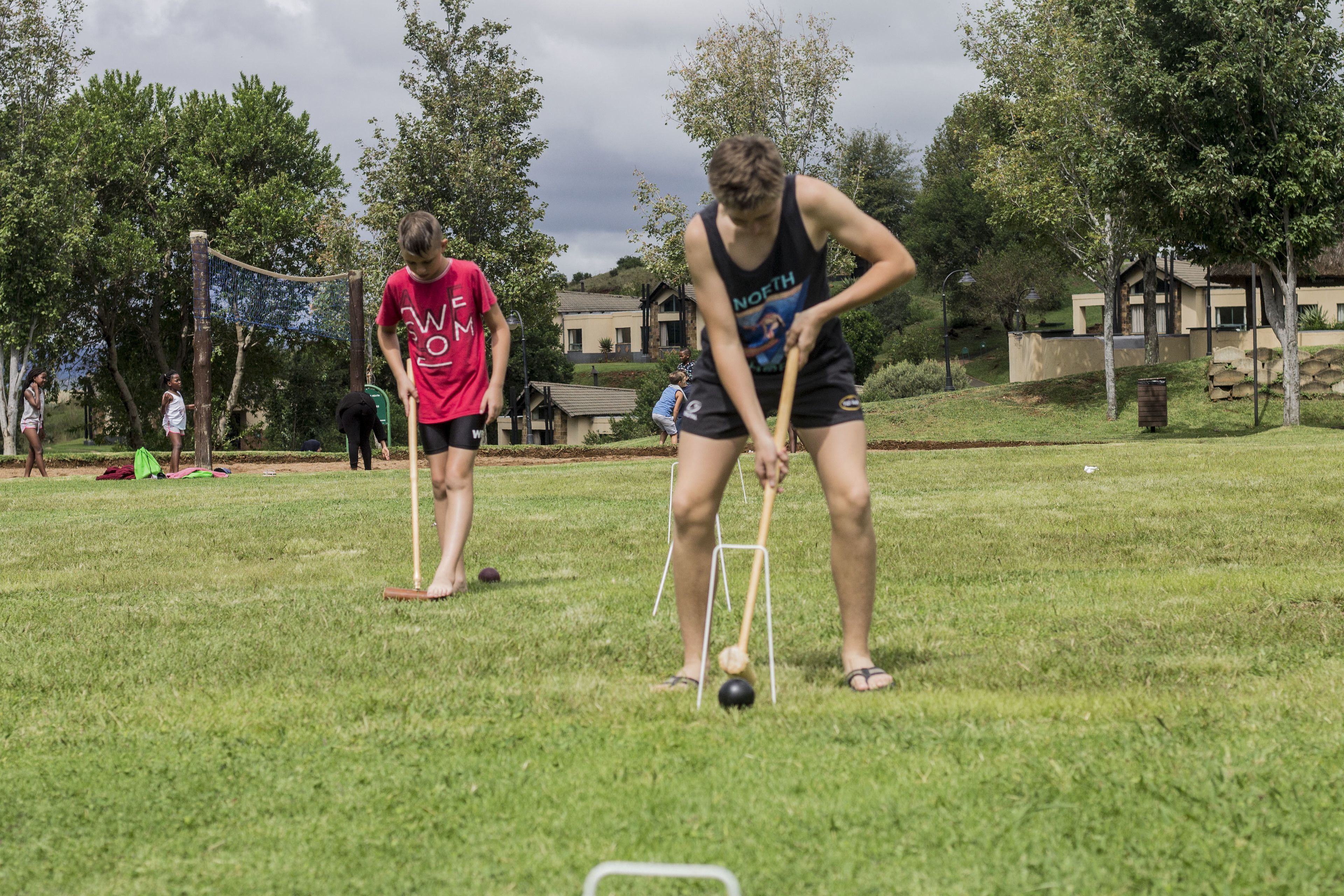 Un espacio al aire libre cubierto de césped, donde personas juegan un partido, posiblemente croquet, con una casa y árboles al fondo.