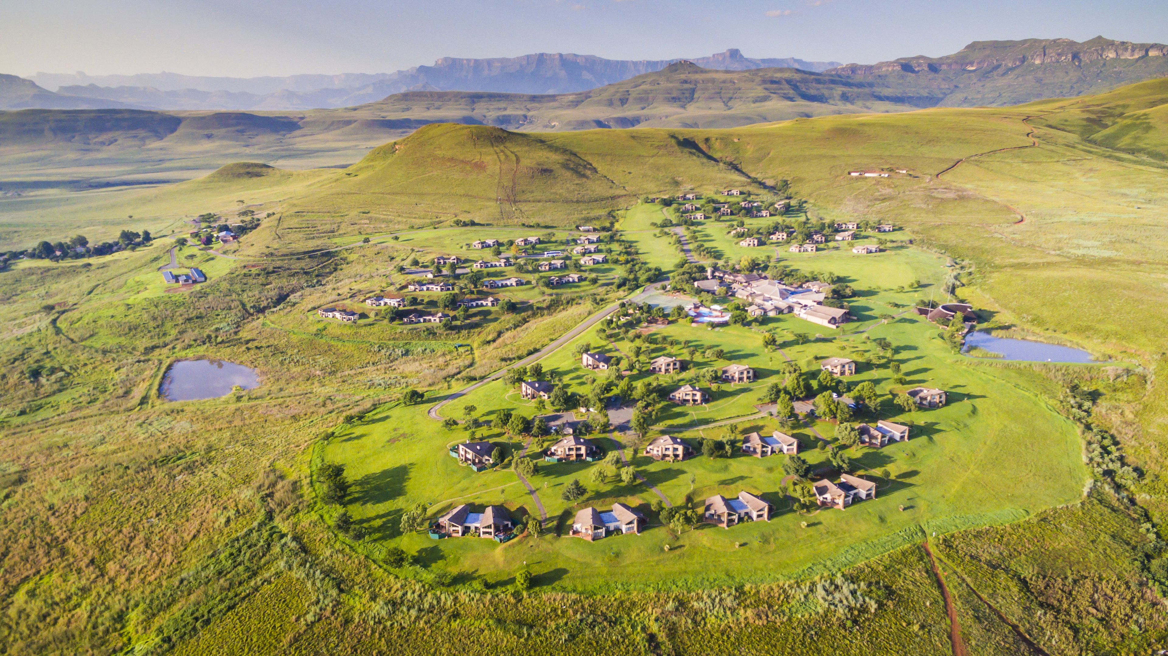 Vista aérea de un área residencial con casas dispuestas en forma de semicírculo alrededor de un espacio verde central, con colinas ondulantes y montañas al fondo.