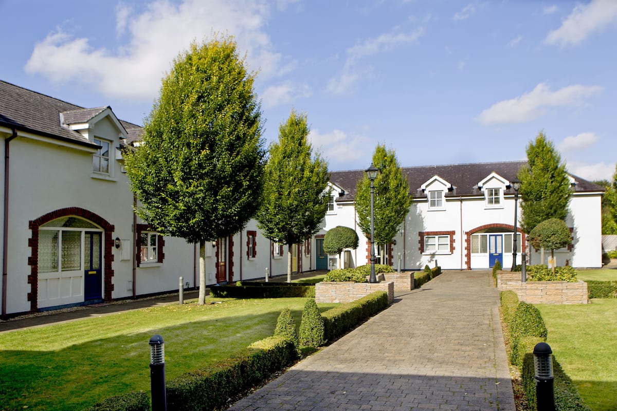 A pathway leading to a row of white buildings with red brick accents, surrounded by well-maintained hedges and trees.
