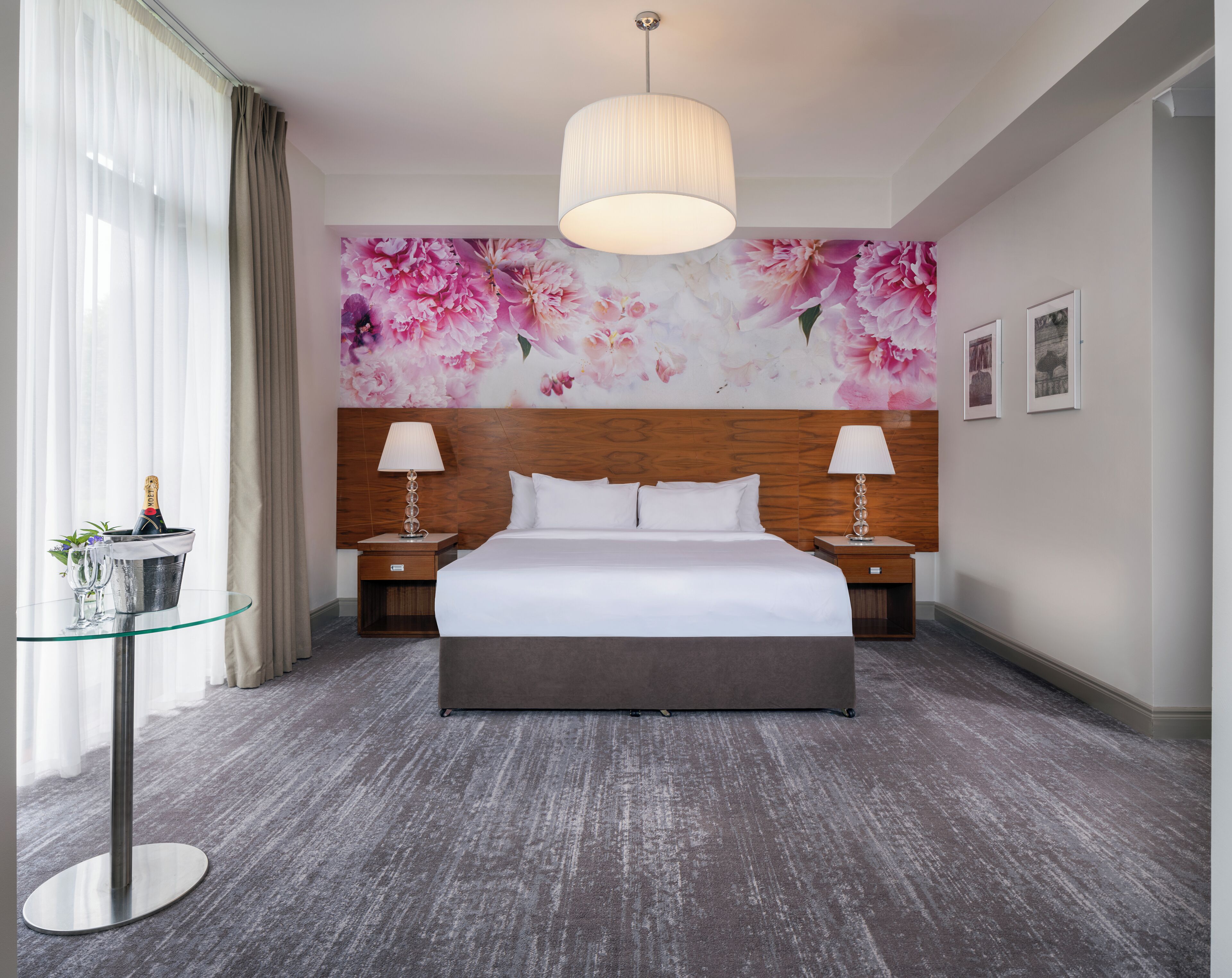 Habitación de hotel con una cama grande, dos mesitas de noche con lámparas, una mesa de vidrio con una botella y vasos, un mural floral en la pared y un candelabro moderno.