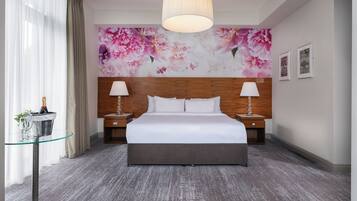 Habitación de hotel con una cama grande, dos mesitas de noche con lámparas, una mesa de vidrio con una botella y vasos, un mural floral en la pared y un candelabro moderno.